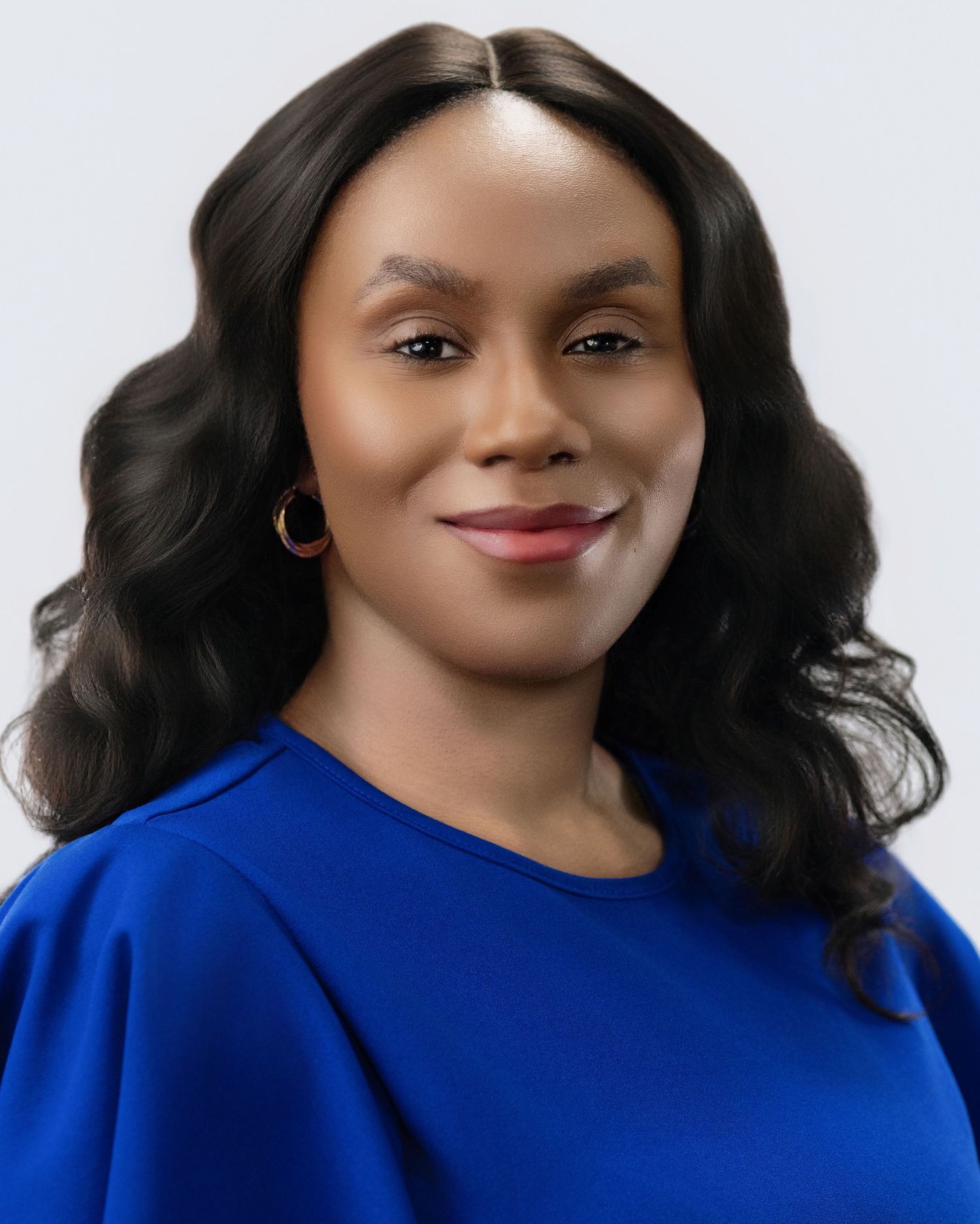 Professional headshot of a smiling woman in a blue top with hoop earrings against a gray background.