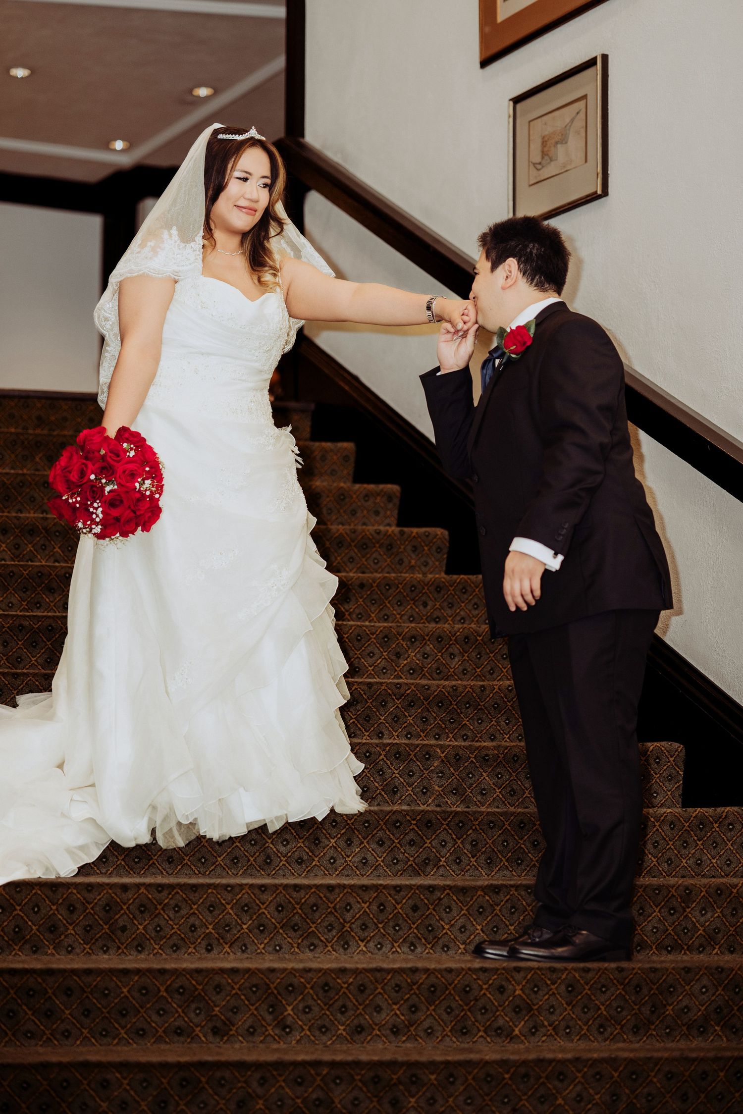 A couple in wedding attire shares a sweet moment on a carpeted staircase, showcasing a white dress and black suit.