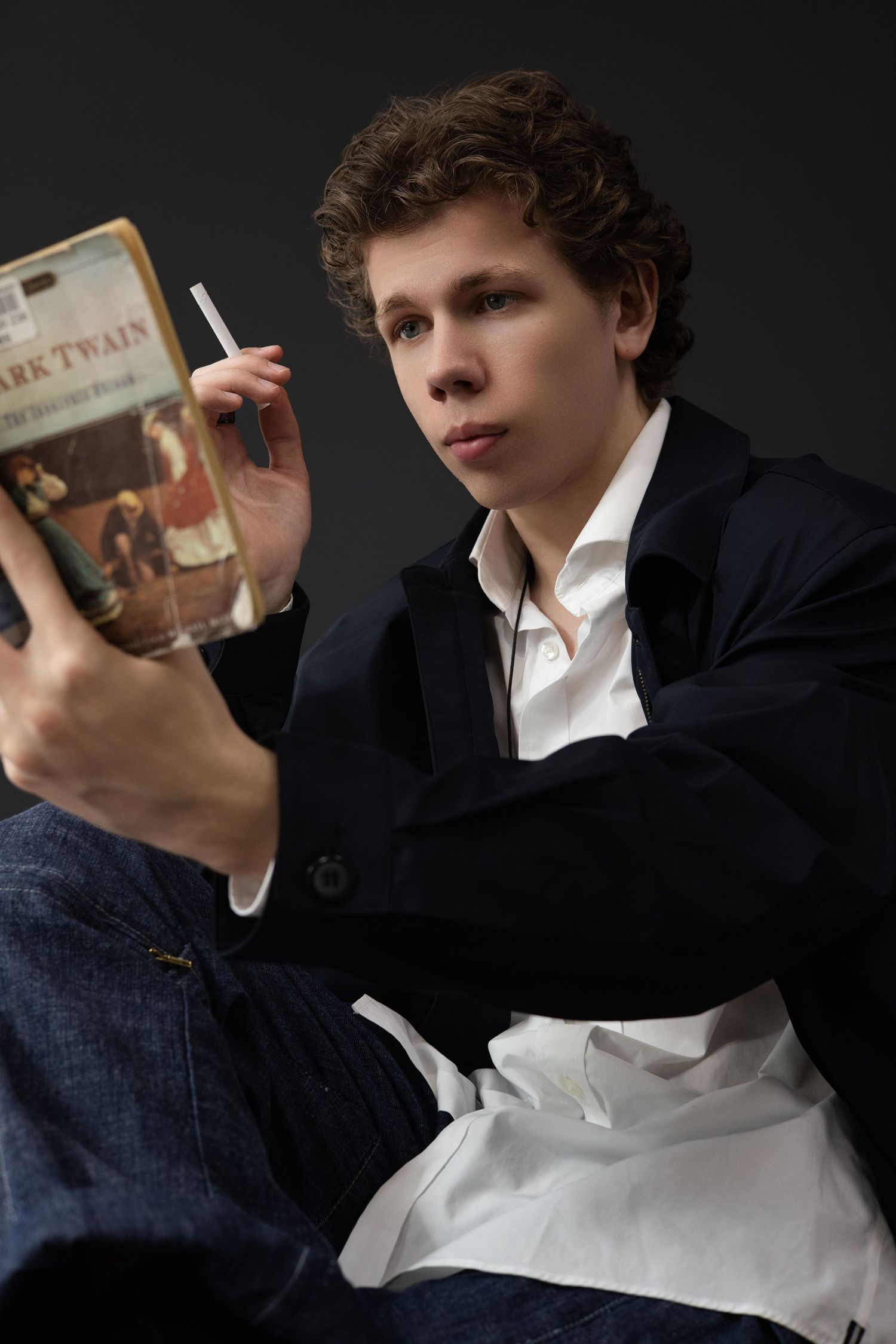 Person in dark suit and white shirt holds book while sitting in dramatic side lighting against dark background.
