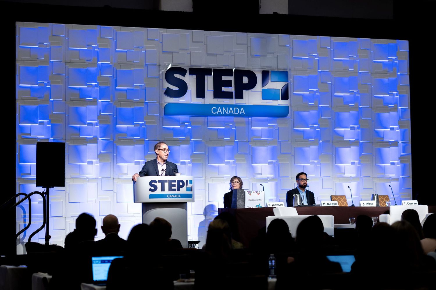 Speaker at STEP Canada conference podium on stage with blue backdrop and audience silhouettes in foreground.
