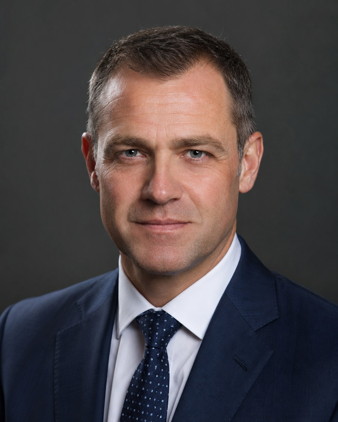 Professional headshot of a man in a navy suit and dotted tie against a dark gray background.