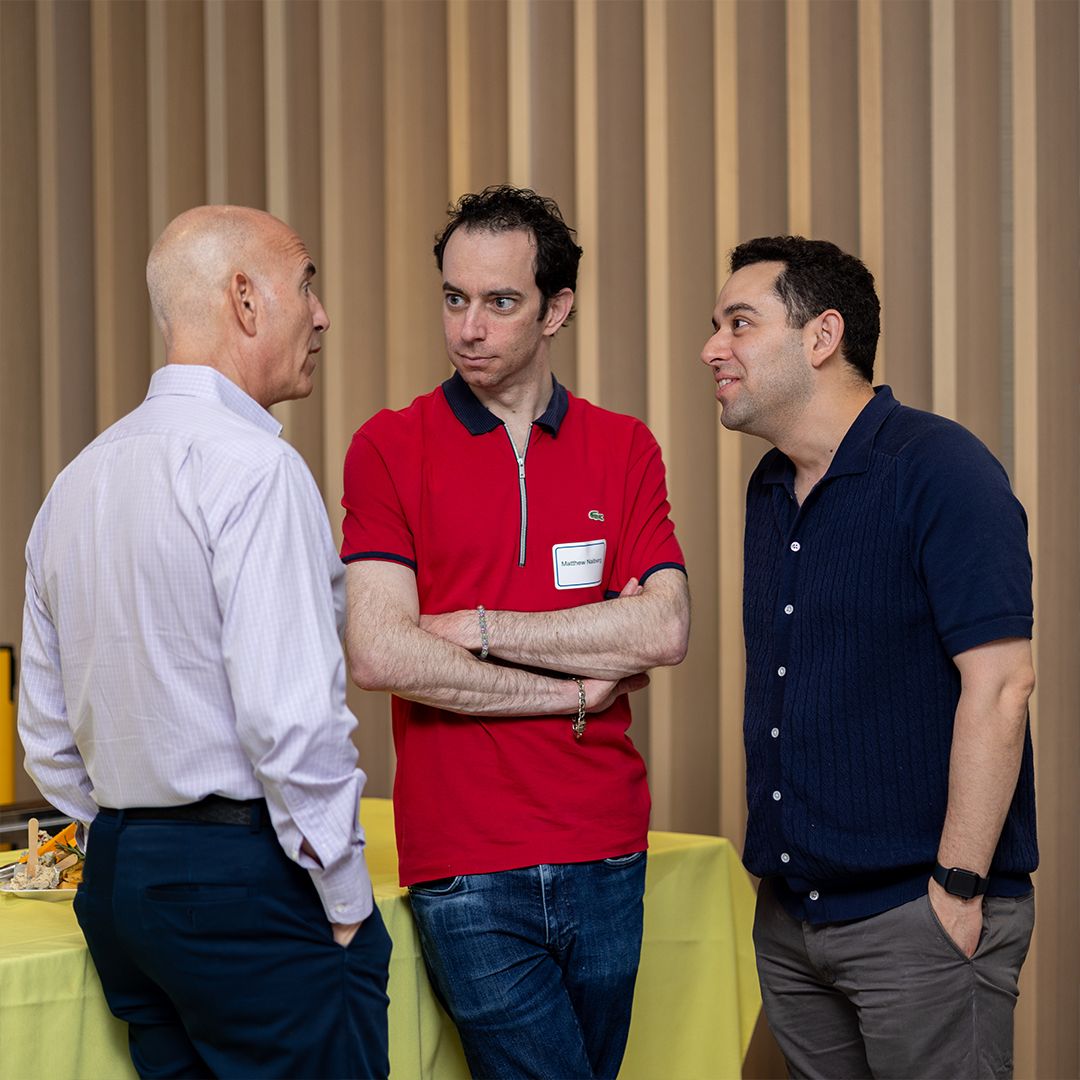 Three men engaged in conversation at a professional networking event, one in red polo, one in navy shirt.