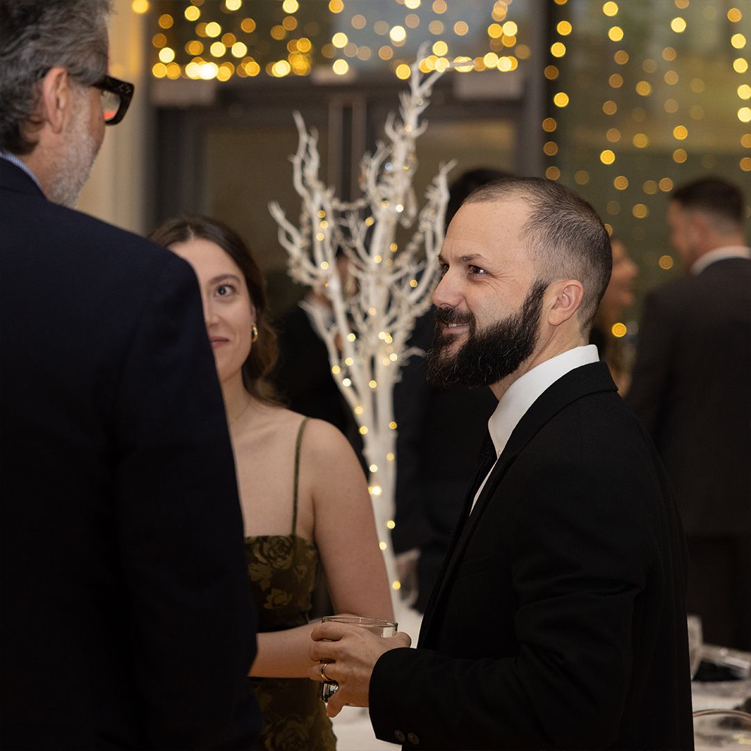 Man in tuxedo with beard conversing with guests at a formal gala event with white tree decorations and string lights.