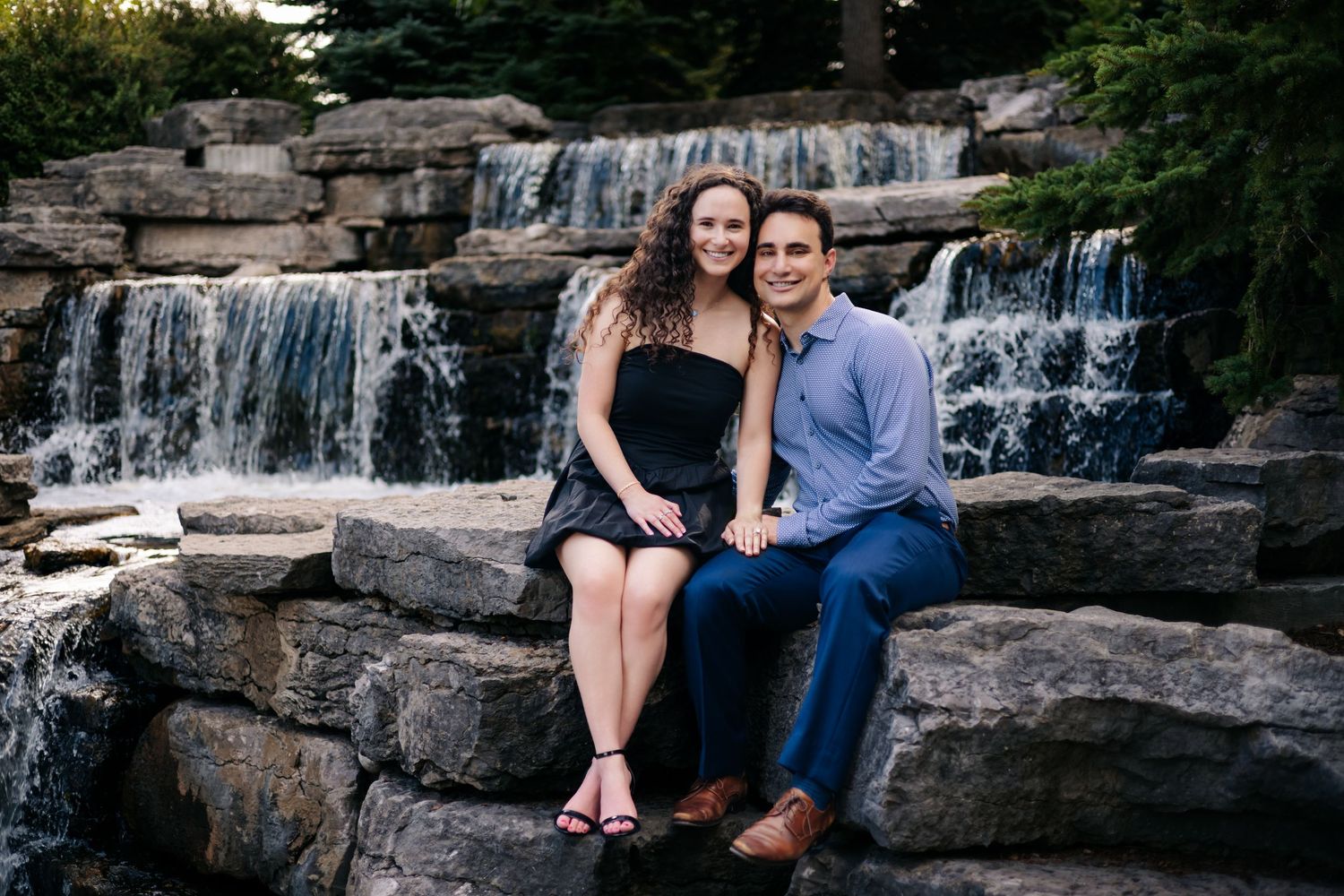 A couple sits together on stone steps beside a cascading waterfall in a scenic outdoor setting.