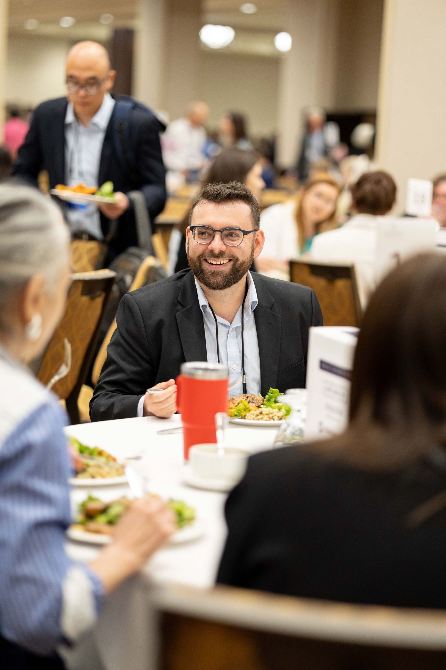 Smiling bearded man in glasses and blazer enjoys a salad lunch at a busy corporate dining event.