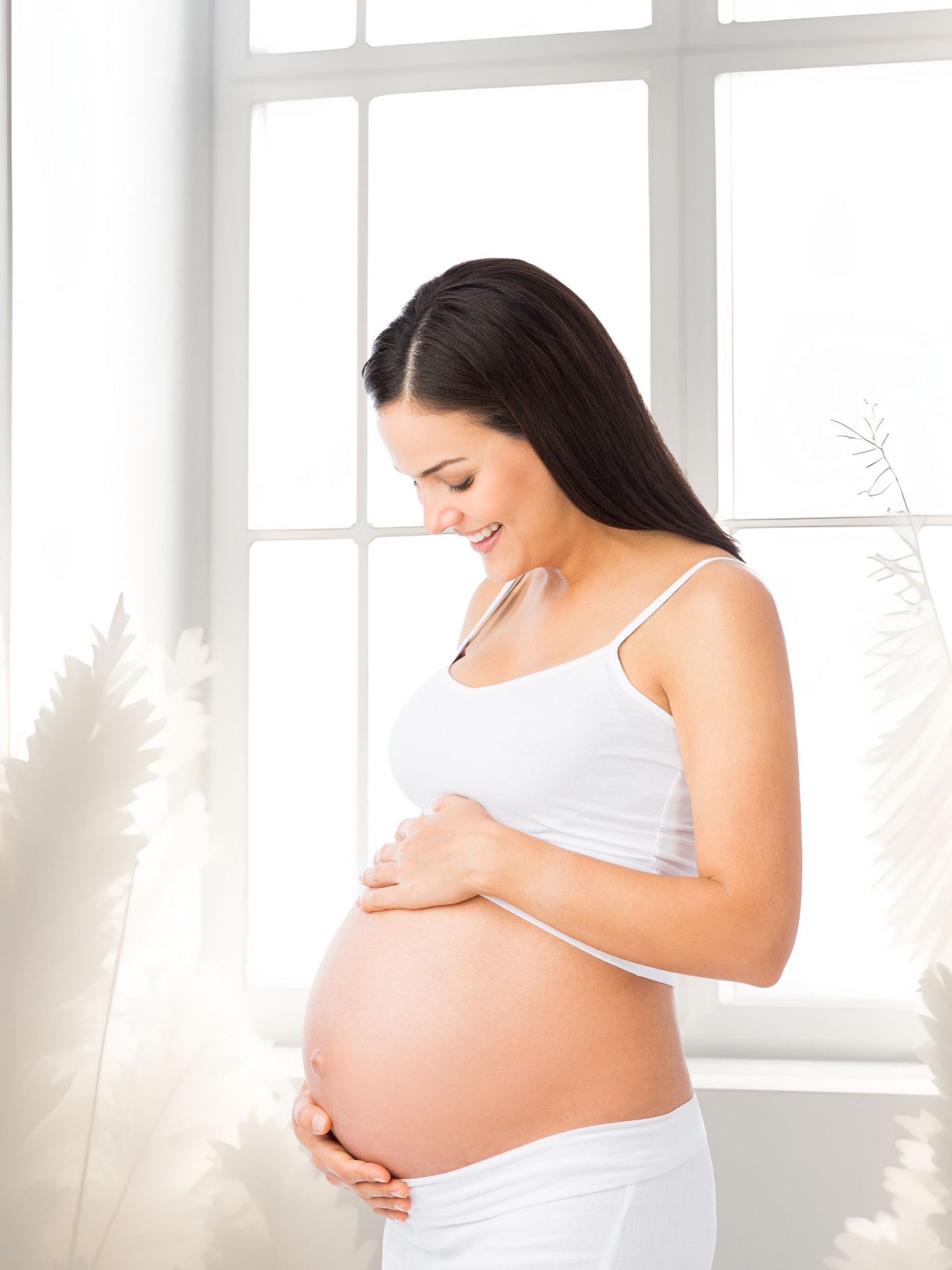 Expectant woman in white tank top caresses her pregnant belly in sunny room with bright windows.