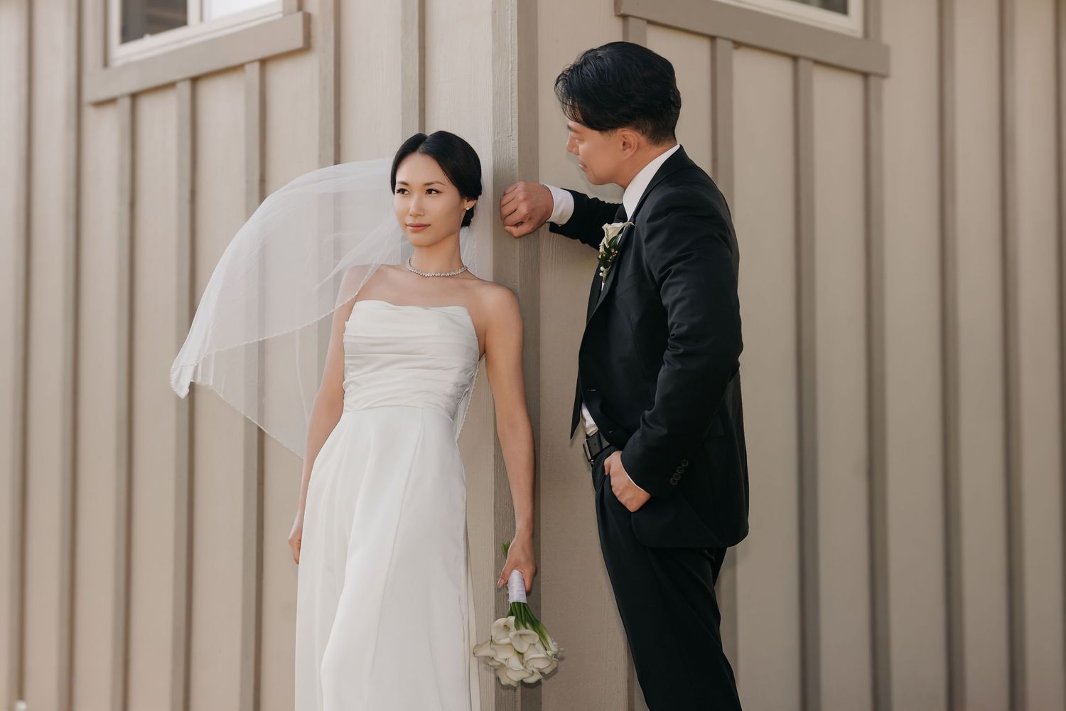 Bride in white strapless gown with veil holds bouquet as groom in black suit adjusts her dress near a wooden wall.