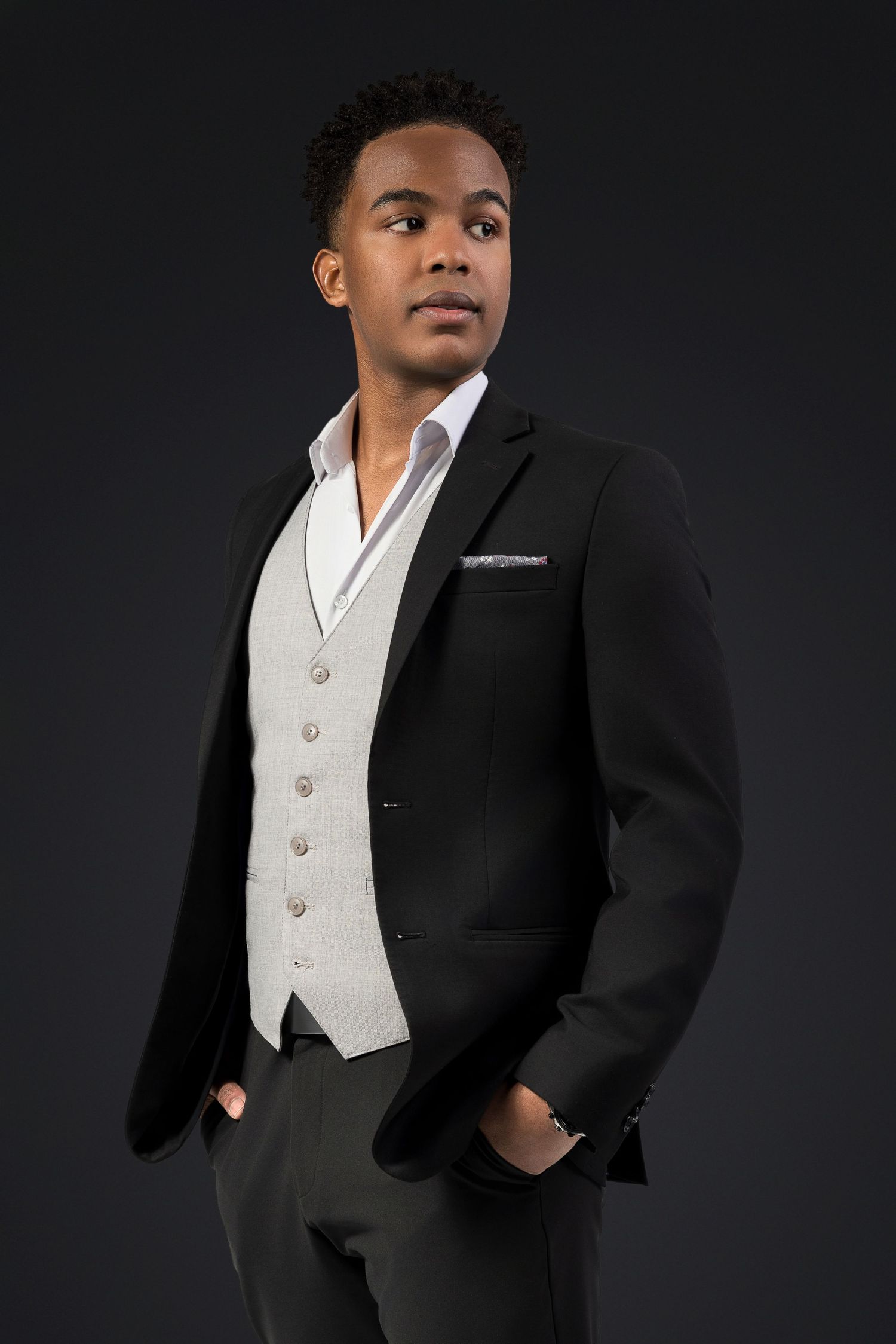 Young person wearing formal black suit jacket and white vest stands elegantly against dark studio background in professional poses.