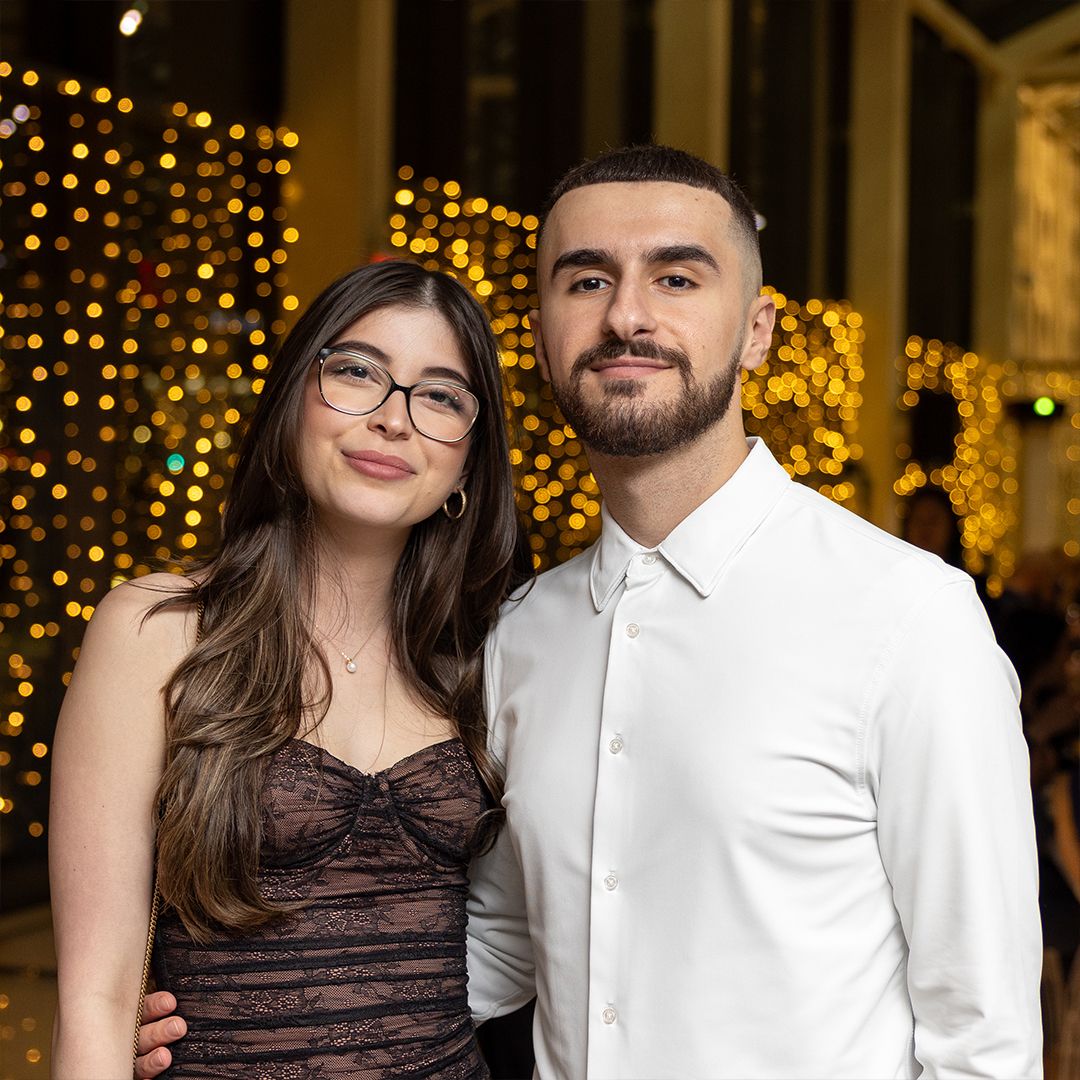 A young couple posing together in front of warm bokeh lights, woman wearing glasses and lace dress, man in white shirt.