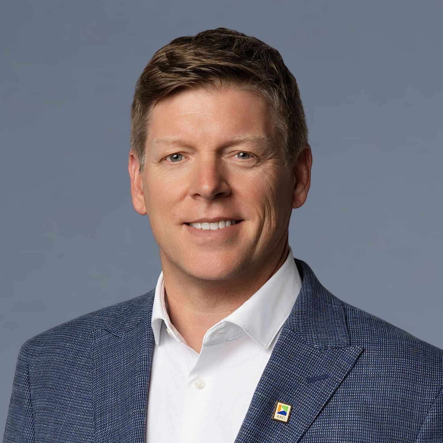 Professional headshot of a smiling middle-aged man in a blue blazer and white shirt against a gray background.