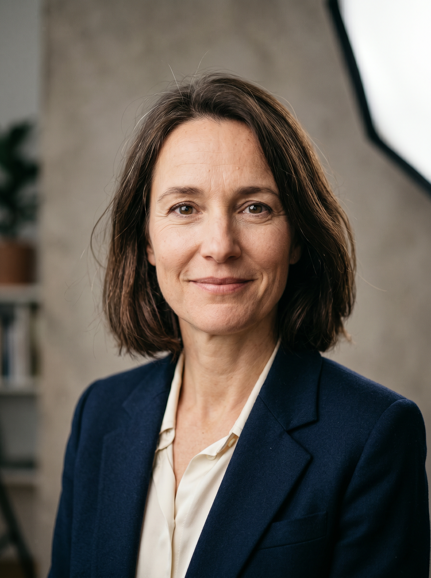 Professional headshot of a woman in a navy blazer with short brown hair, smiling confidently in a modern studio setting.