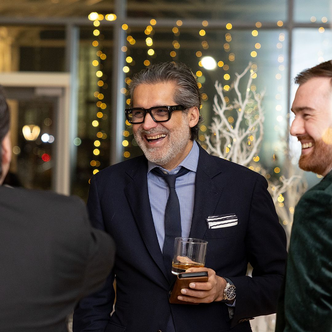 Two men in suits laughing and socializing at an elegant evening corporate event with warm bokeh lights in background.