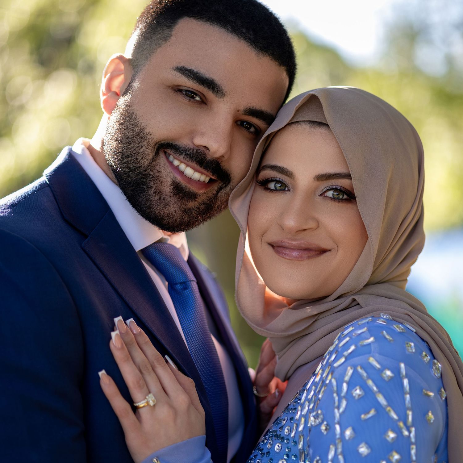 A couple in formal attire poses together outdoors with the person on the right wearing a blue floral dress and hijab.