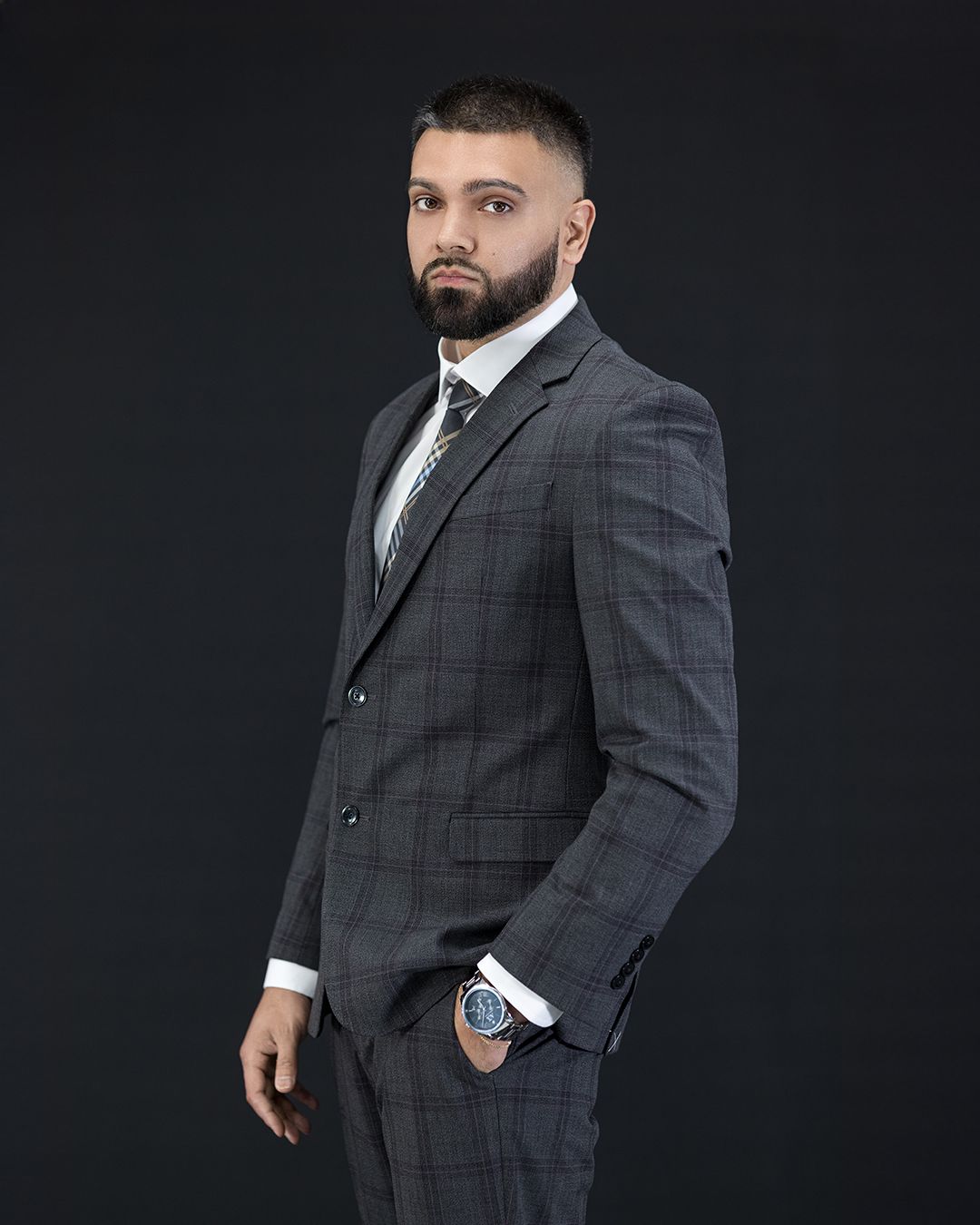 Professional studio portrait of a figure in a well-fitted gray plaid suit and dark tie against a black background.