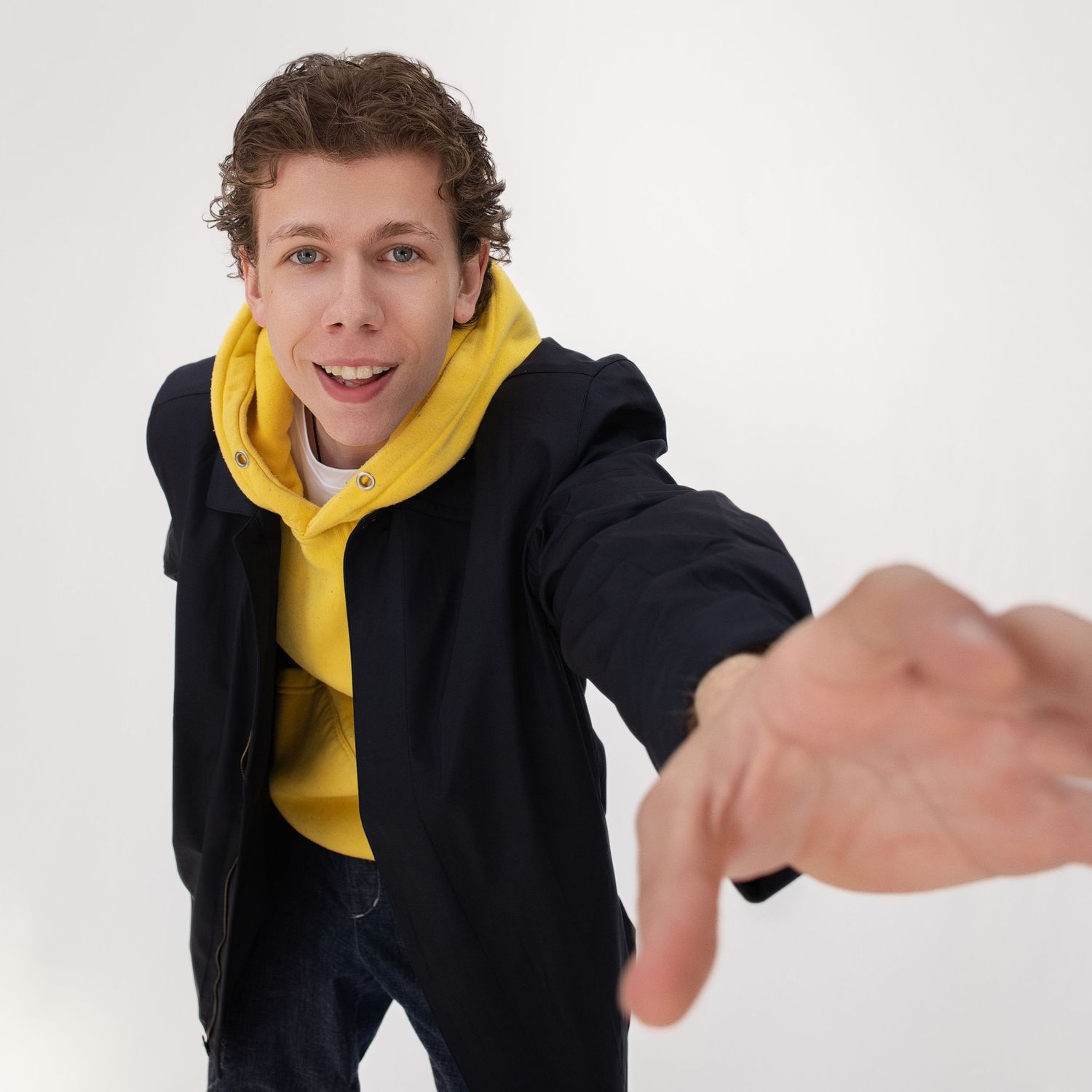 Person wearing black jacket and yellow hoodie strikes playful pose against white backdrop.