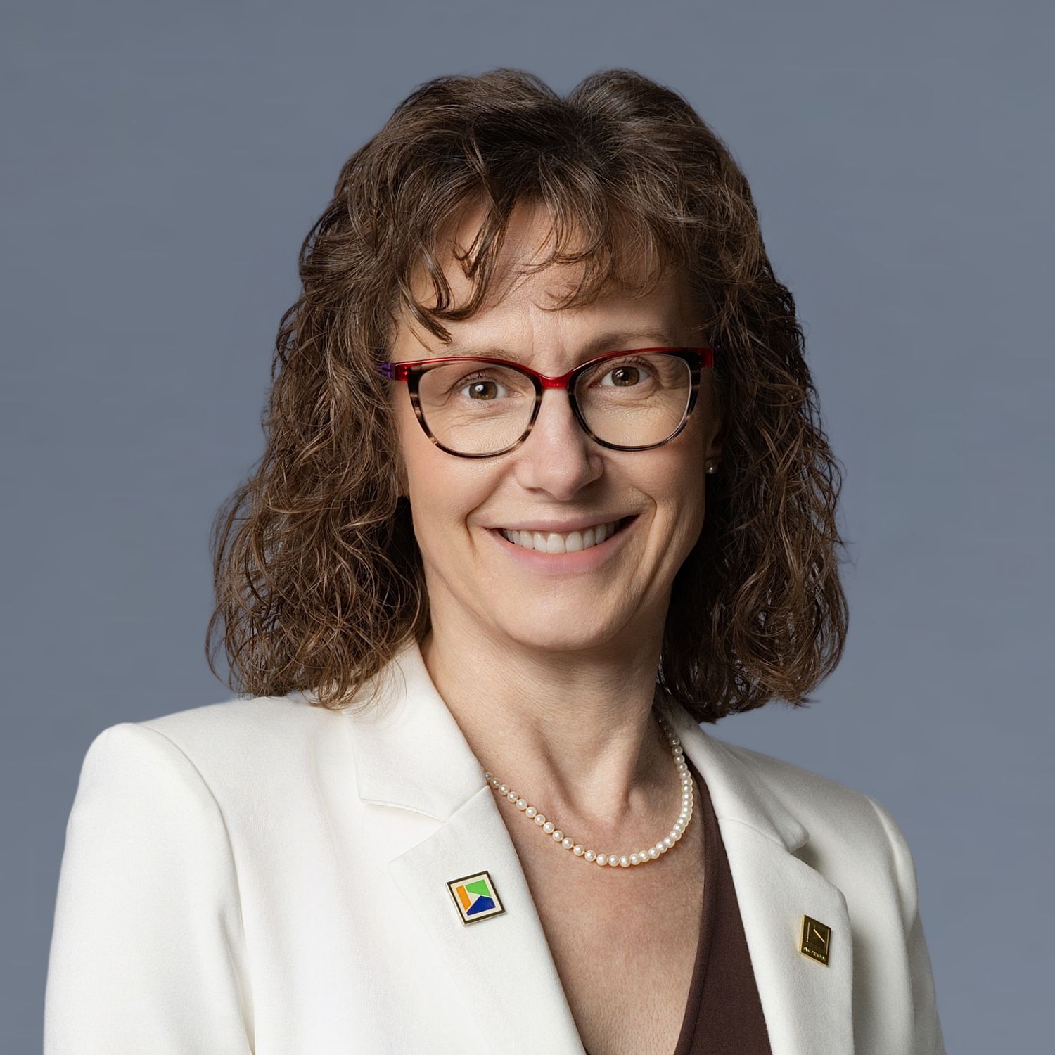Professional headshot of a smiling woman with curly brown hair, red glasses, pearl necklace, and white blazer on gray background.