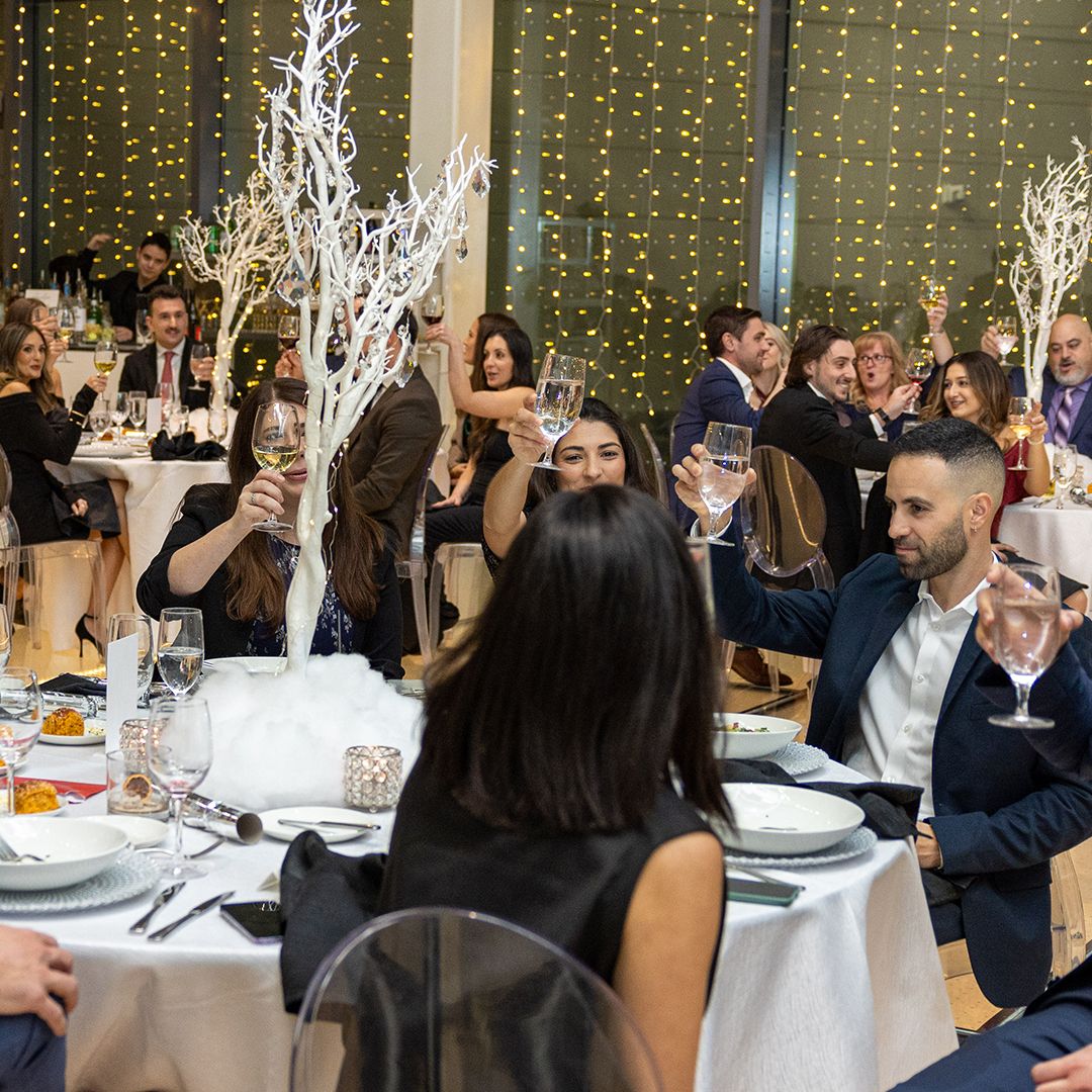 Elegantly dressed guests raising champagne glasses at a formal banquet dinner with white floral centerpieces and fairy lights.