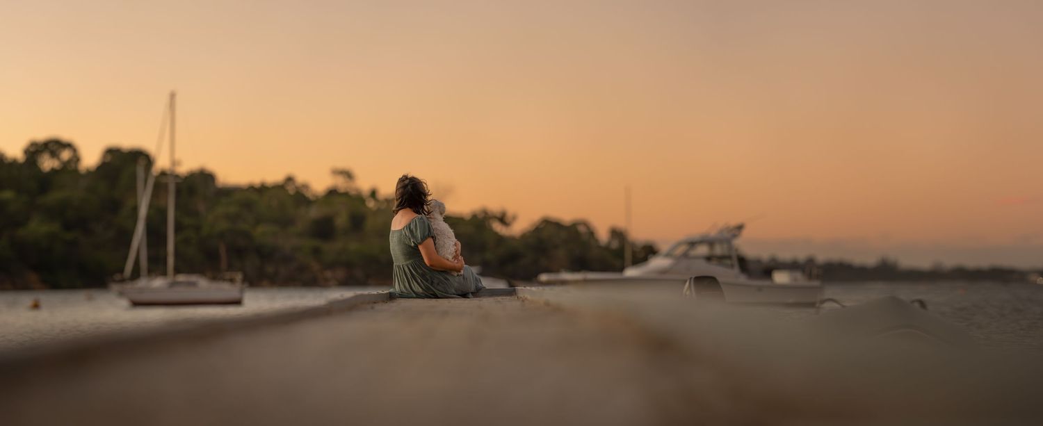 A girl and her dog sitting on a pier at sunset time looking across the river. You can see water and boats to the side.