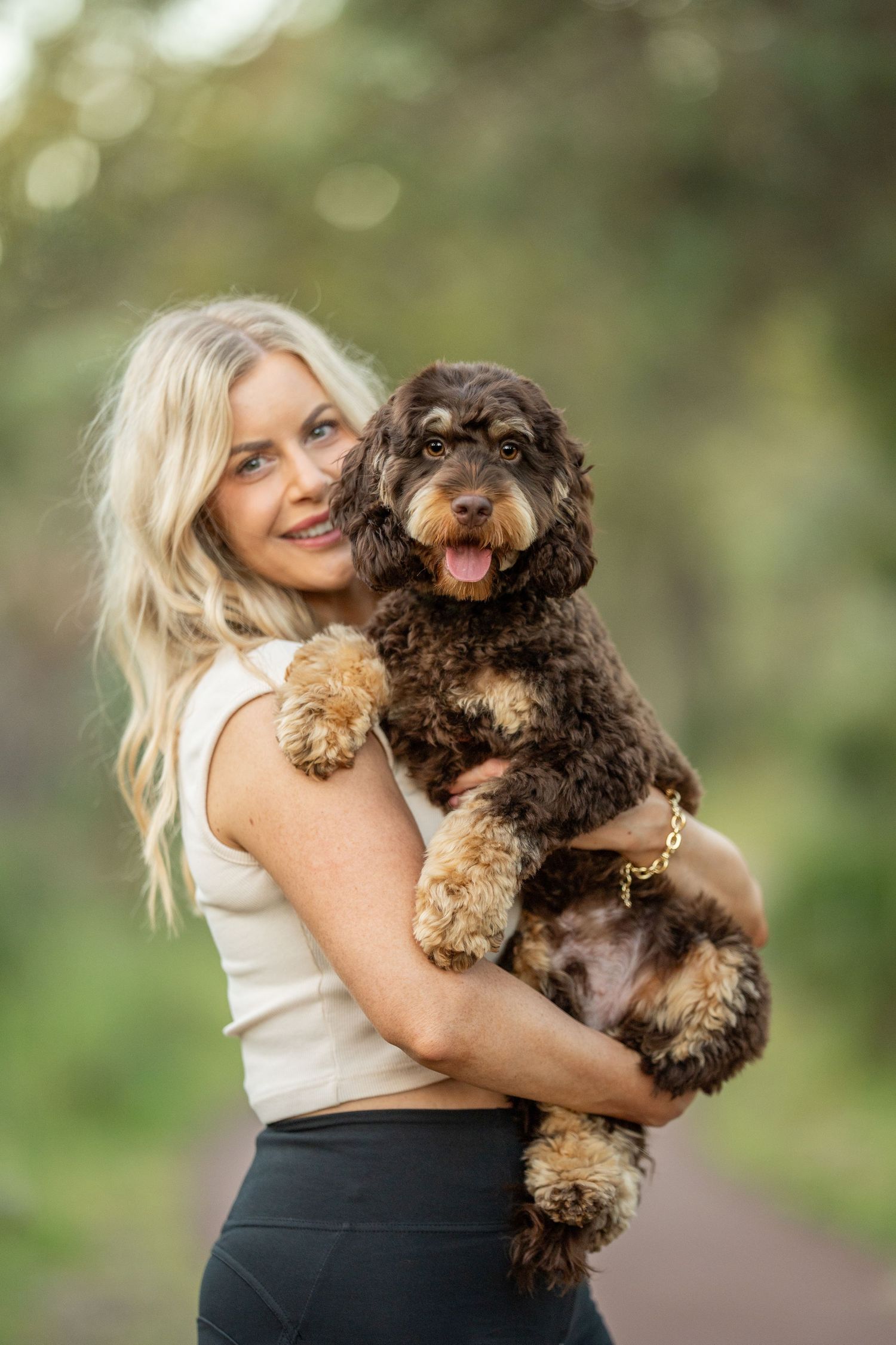 A woman in black pants and white top holds and cuddles an adorable brown cockapoo puppy outdoors in nature.