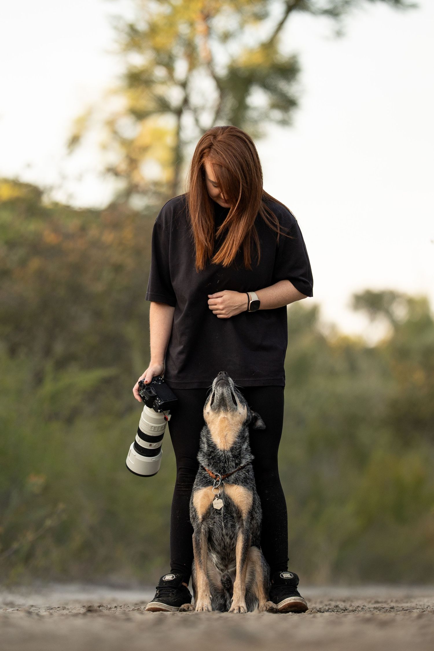 Photographer in black outfit stands with camera gear alongside attentive Australian Shepherd dog on dirt path at sunset.