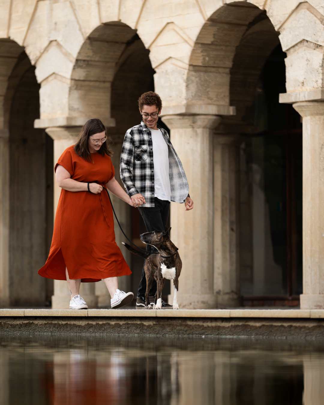 A couple stands with their dog under stone archways near a reflective body of water on a sunny day.