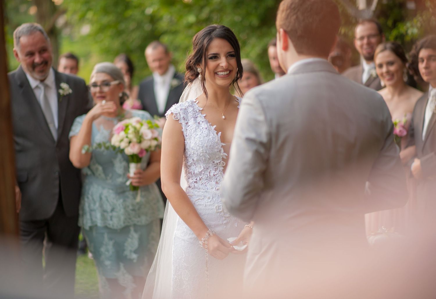 A wedding ceremony taking place outdoors with a bride in a white lace dress holding hands with someone during the vows.