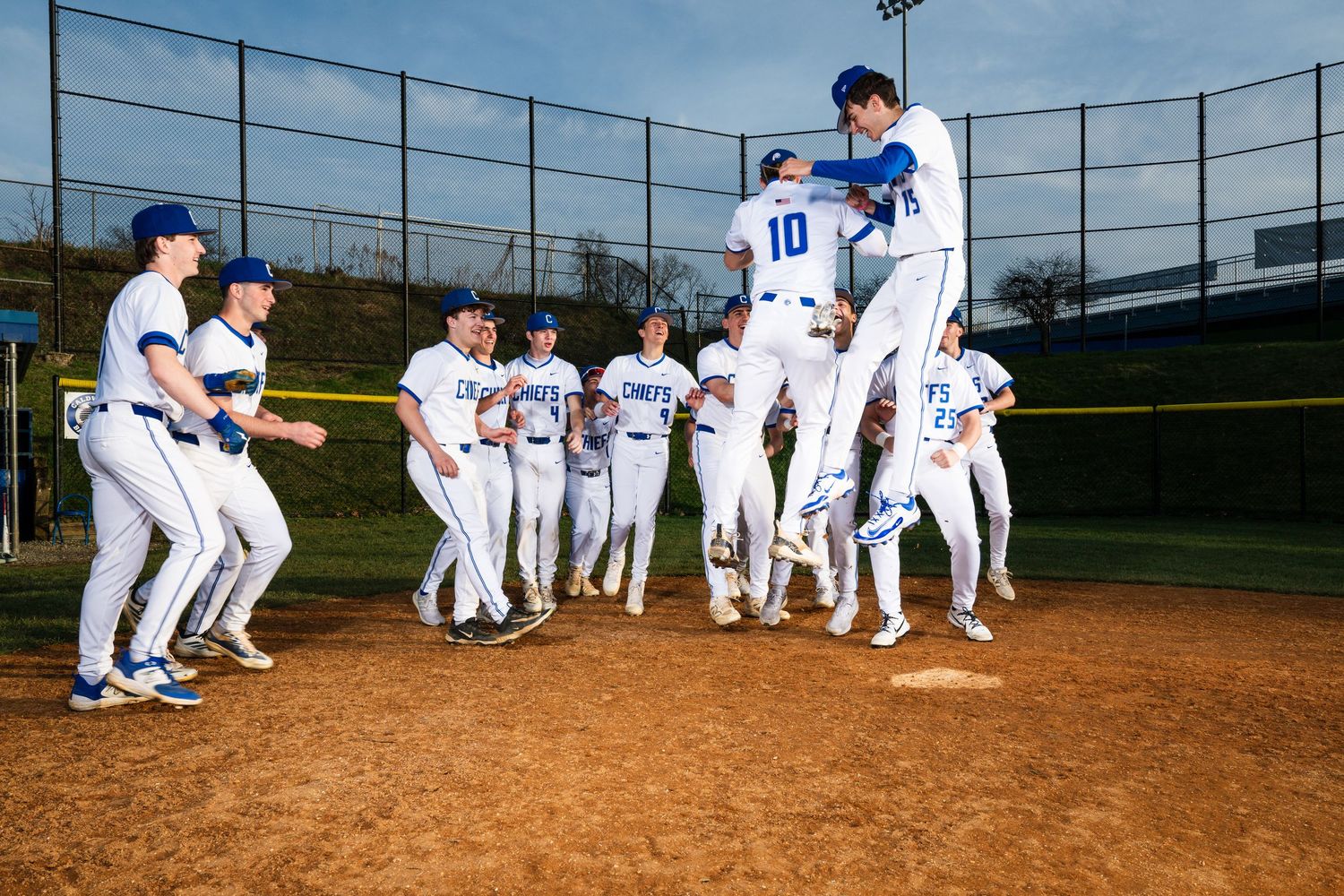 High school baseball team in blue and white uniforms celebrating on the field with players jumping and cheering.