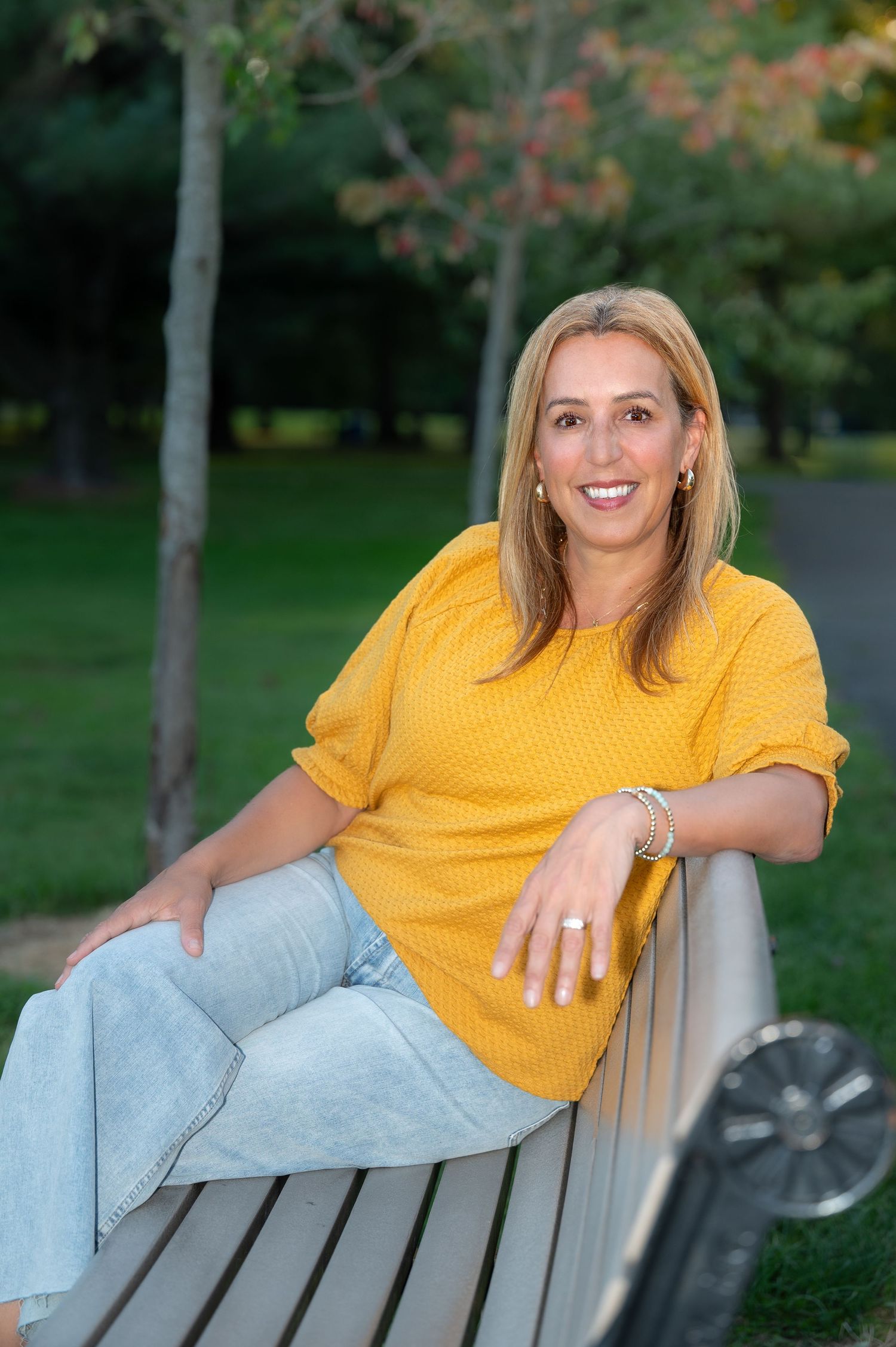 A person in a yellow sweater and light jeans relaxes on a park bench during autumn.
