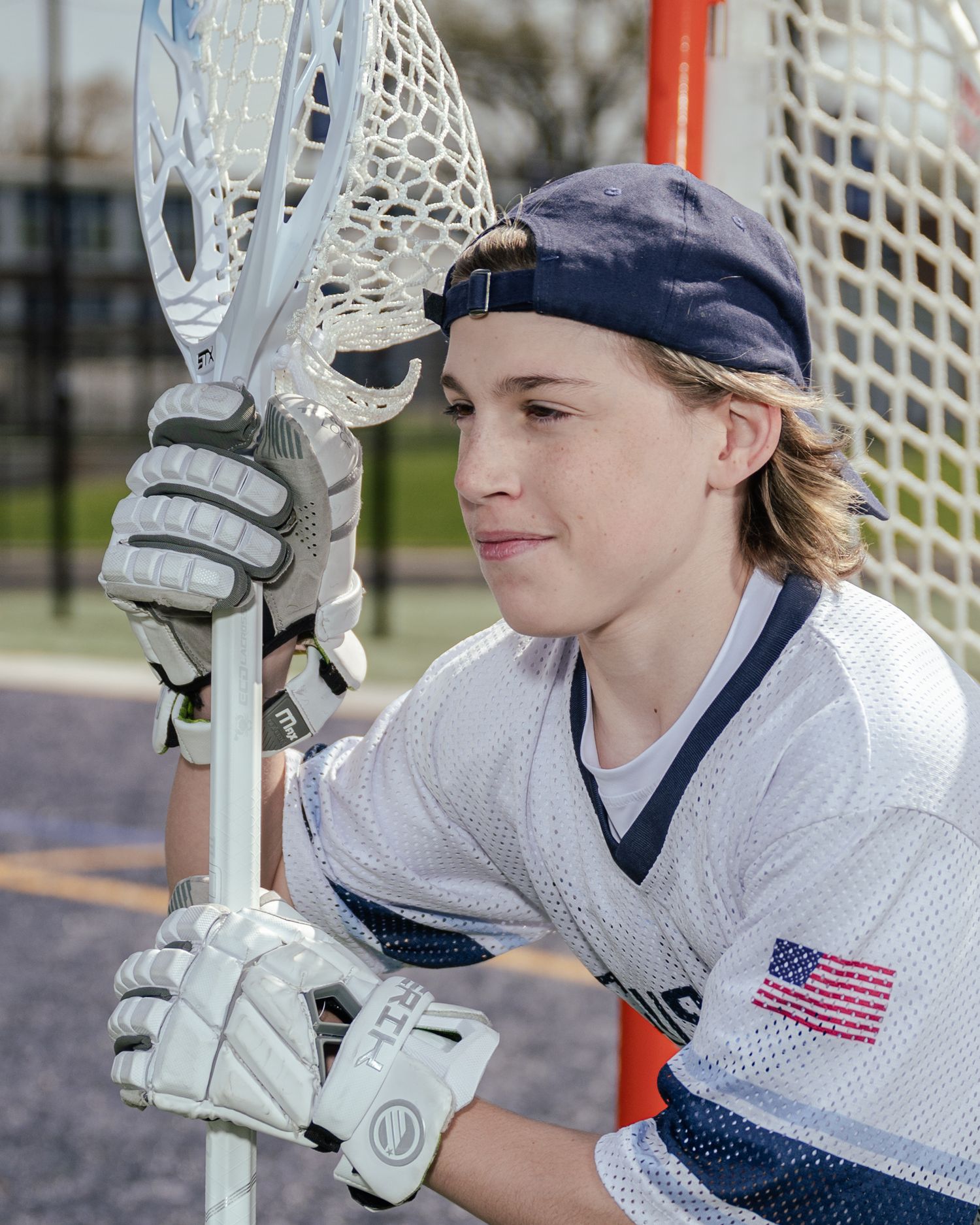 Lacrosse player in white uniform and navy cap poses with equipment by goal net.