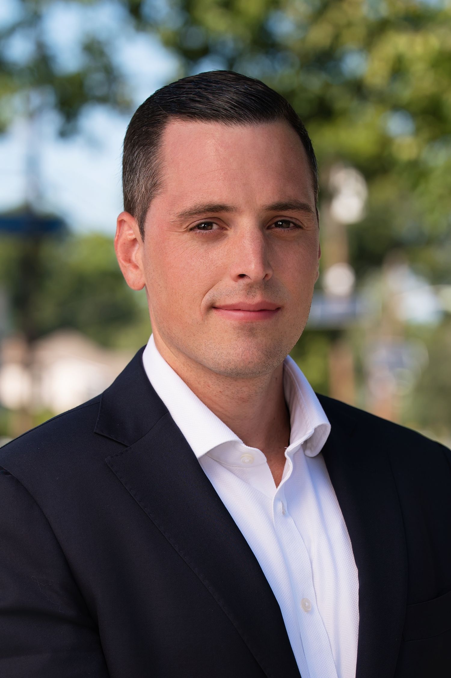 Professional headshot of someone in a dark suit jacket and white dress shirt against a blurred outdoor background.