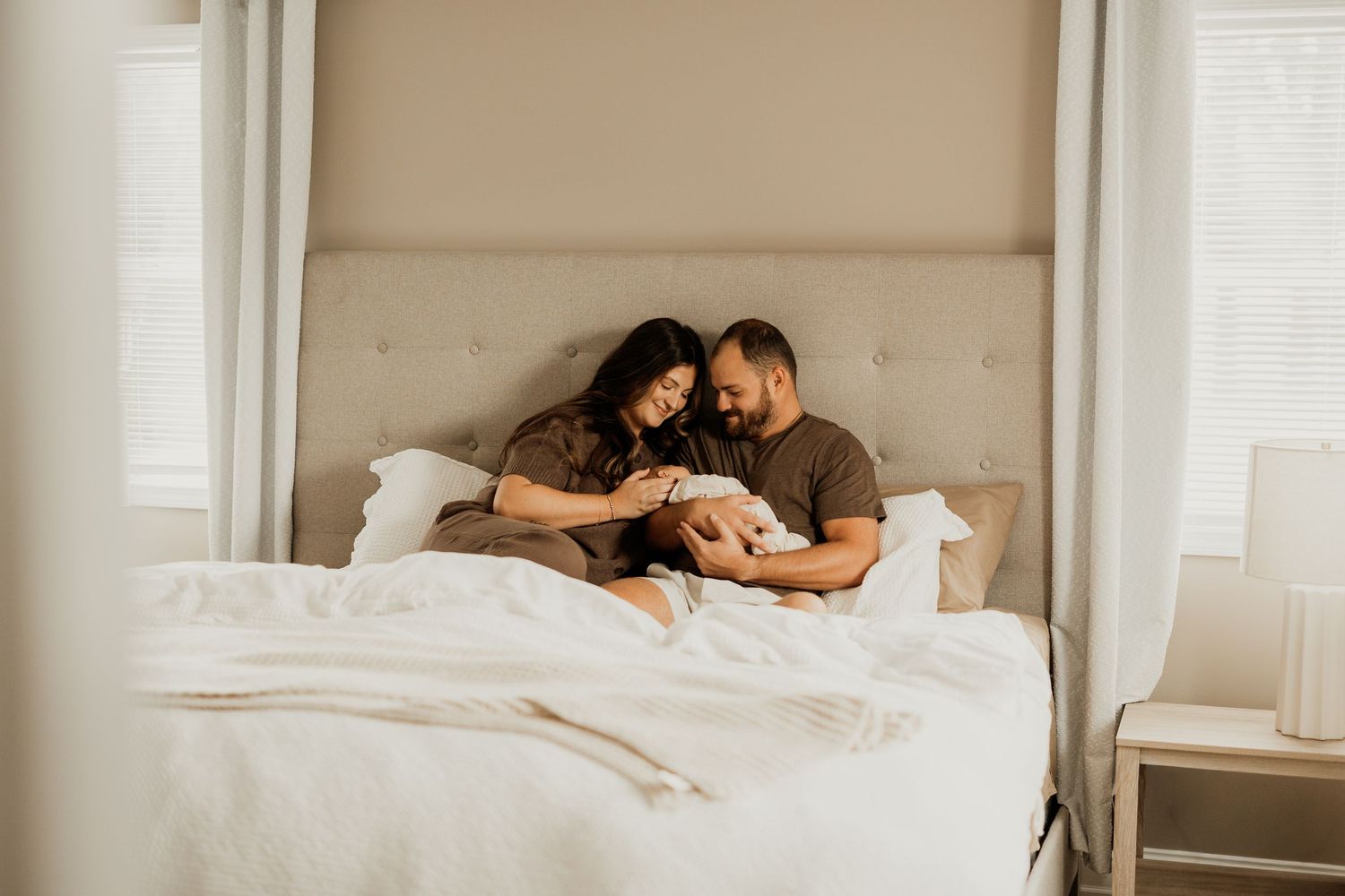 Couple sitting in bed together admiring their newborn baby in a bright, cozy bedroom.