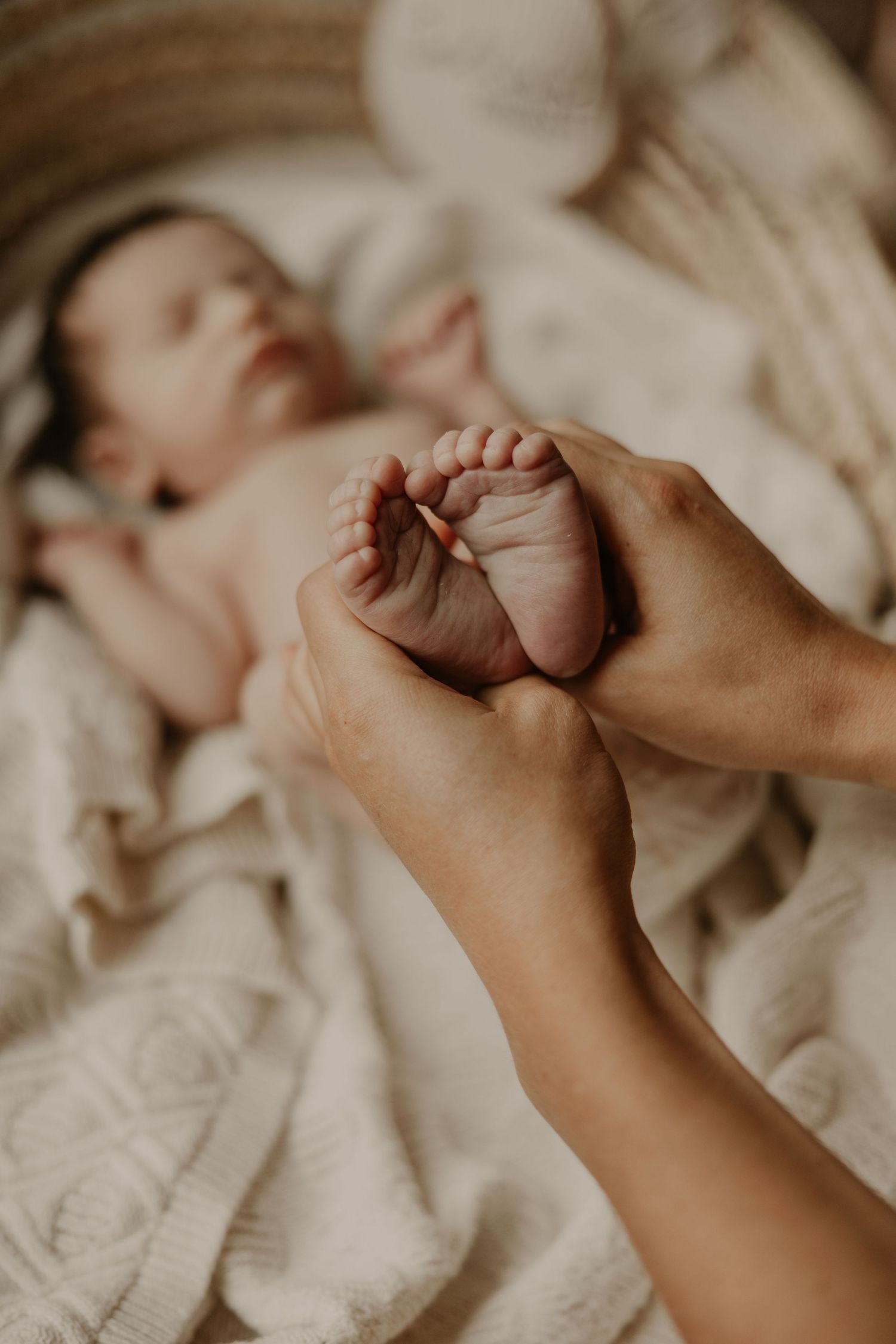 Adult hands gently holding tiny newborn baby feet, with sleeping infant on white blanket in soft natural light.