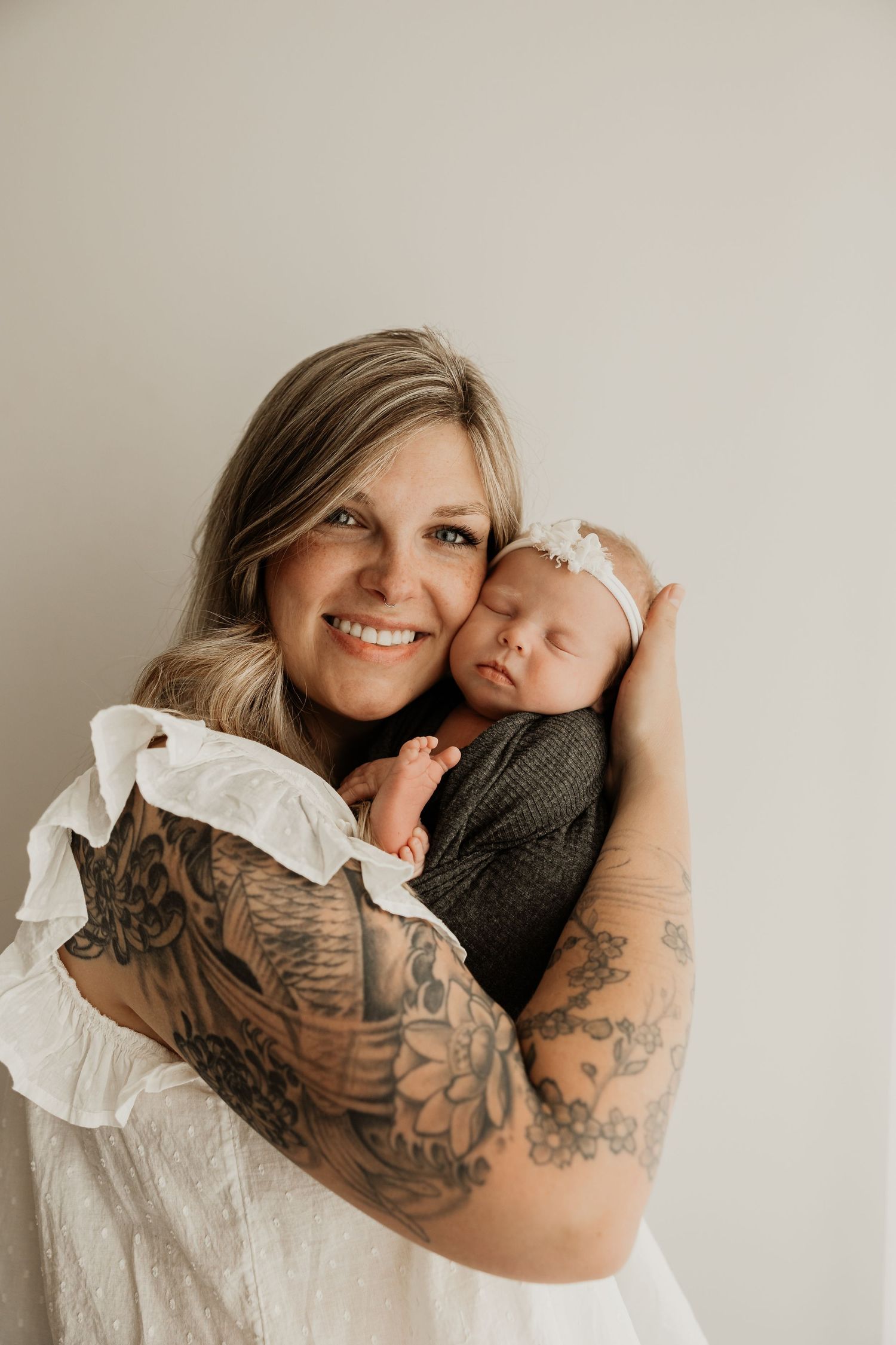 Smiling tattooed mother holding sleeping newborn baby wrapped in dark swaddle with white headband against light background.