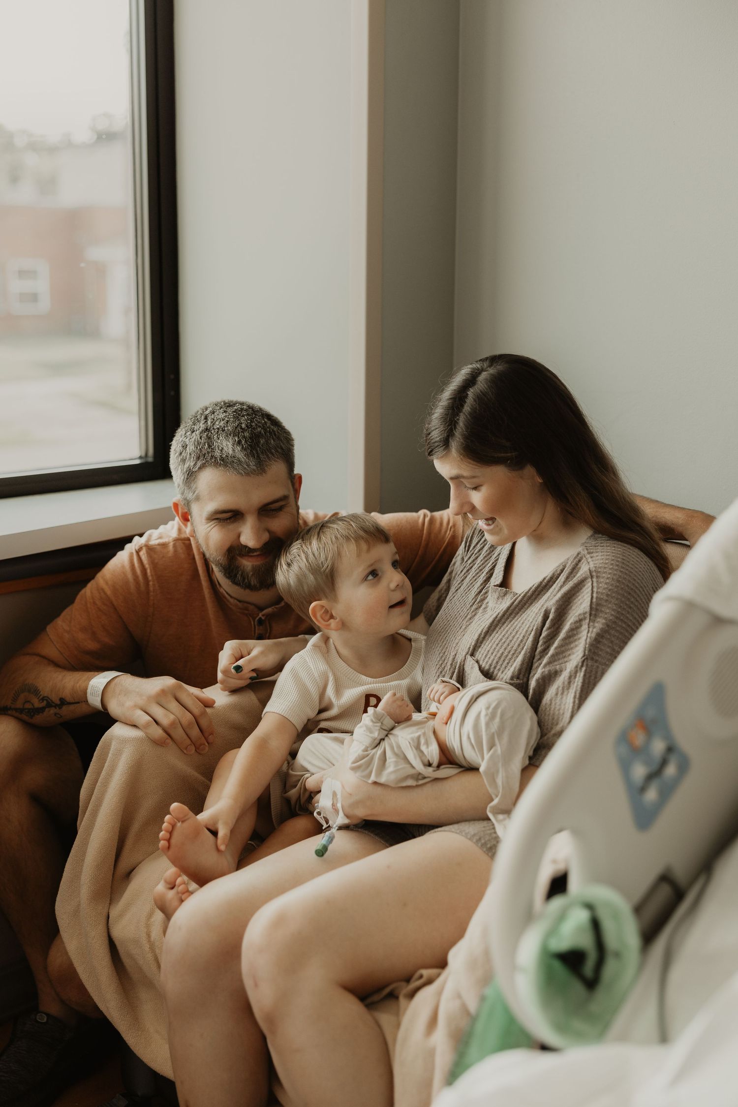 A happy family with a toddler sits together on a couch by a window in a warm, cozy room.