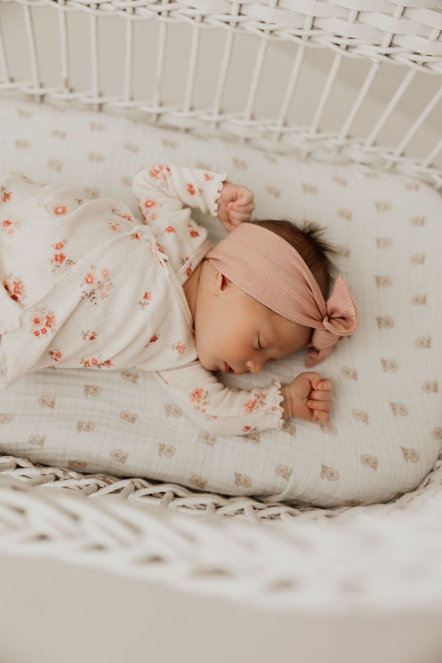 Newborn baby girl sleeping peacefully in a white wicker bassinet, wearing a floral outfit and pink bow headband.