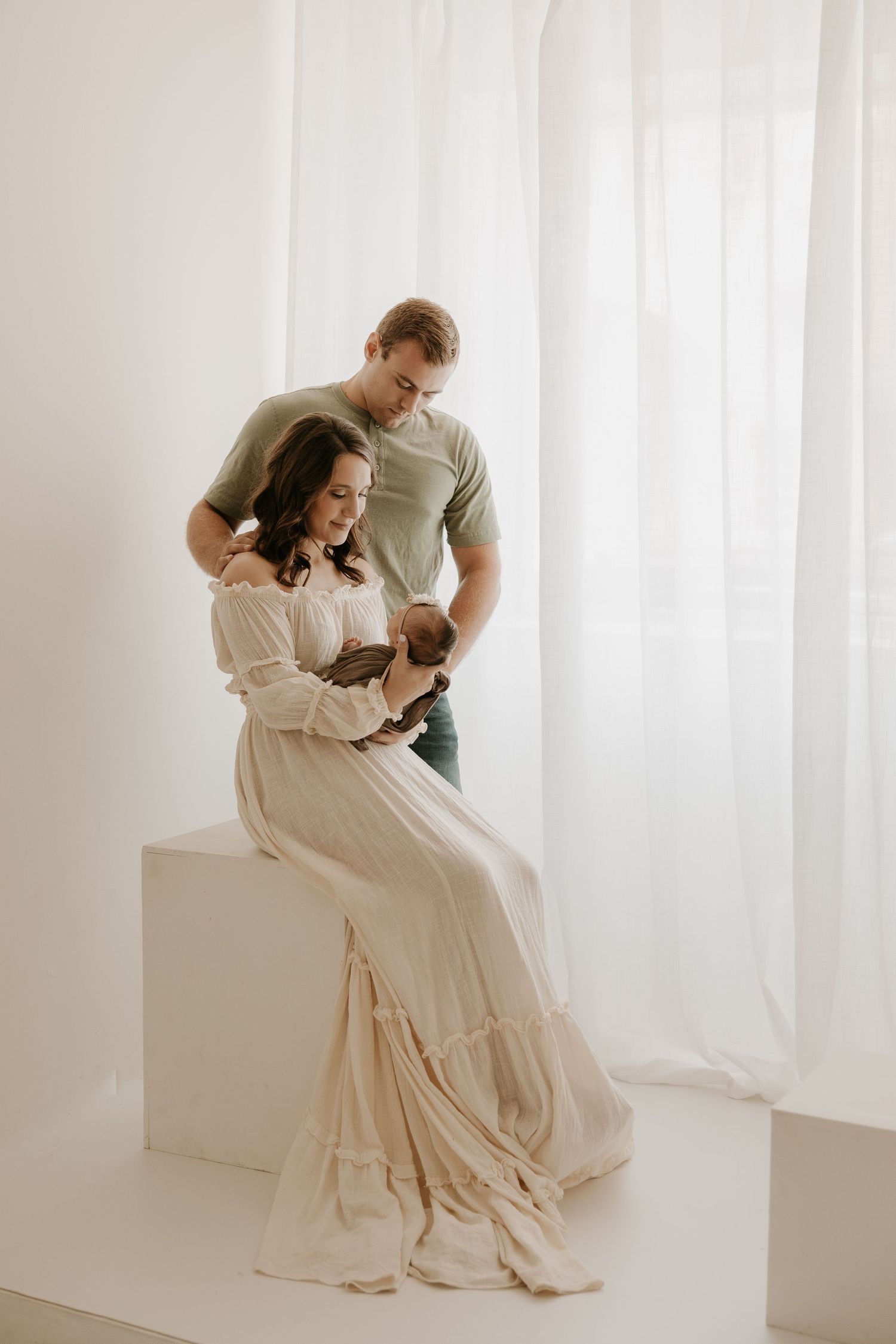 A couple tenderly holds their newborn baby in a bright white studio, the mother in an elegant cream dress.