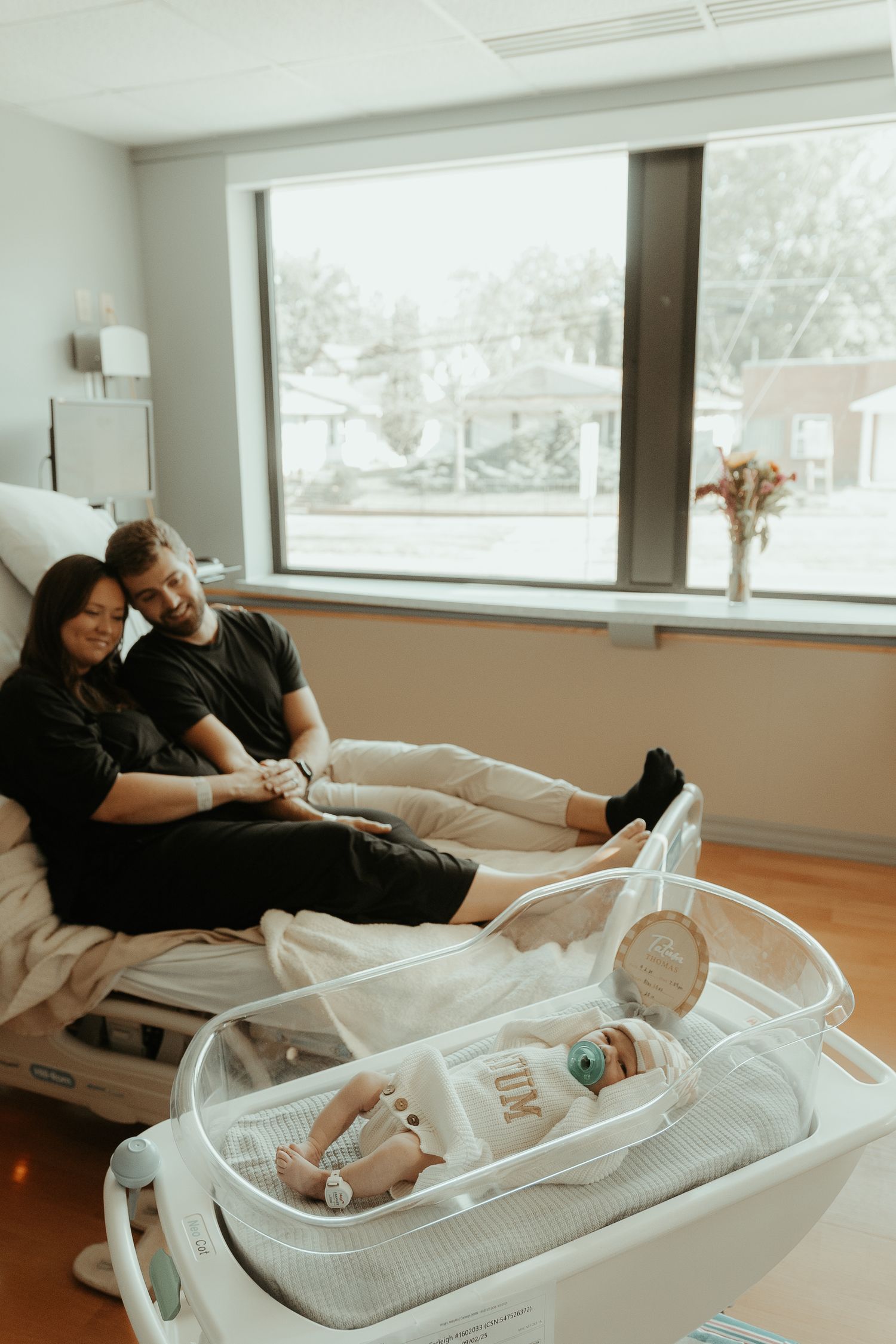 New parents resting on hospital bed smiling at their newborn baby in a clear bassinet after delivery.