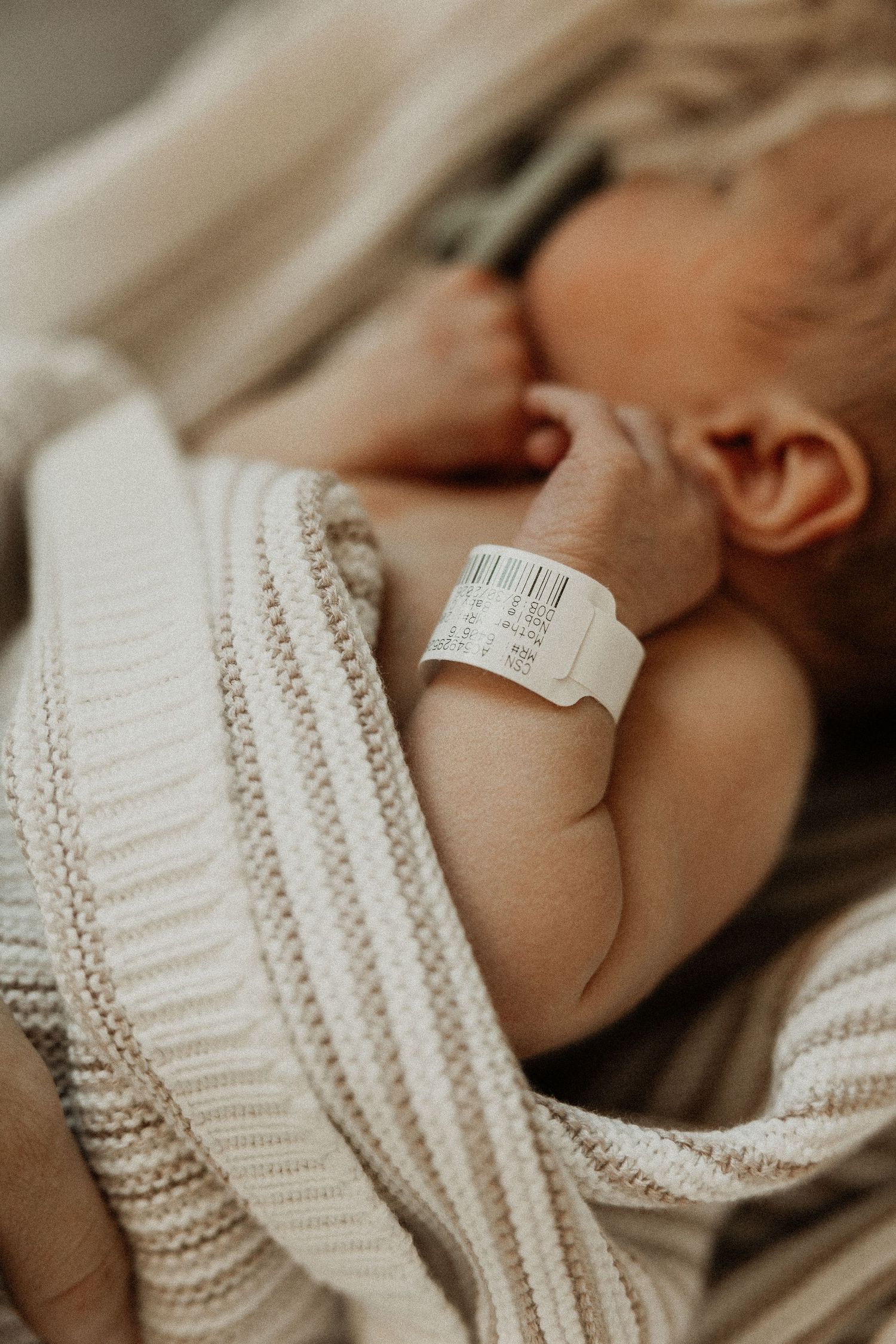 Newborn baby sleeping peacefully wrapped in a cream knit blanket with a hospital identification bracelet on wrist.