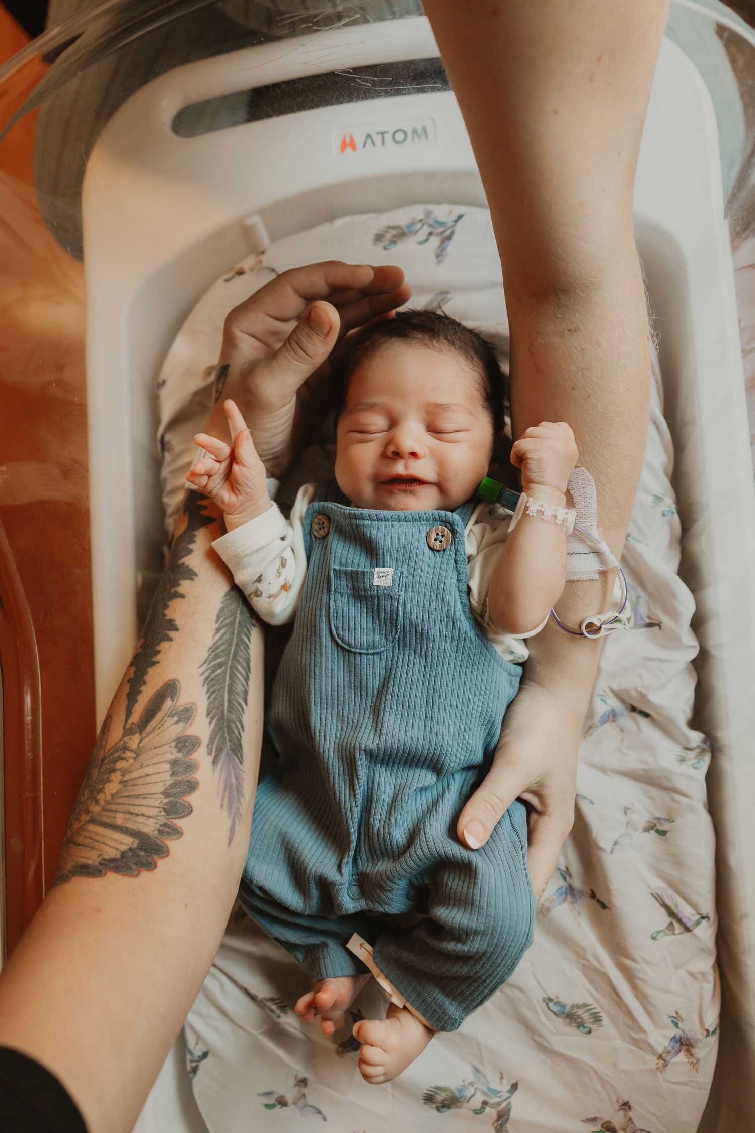 Newborn baby smiling in blue overalls on hospital bassinet, with tattooed parent hands cradling them.