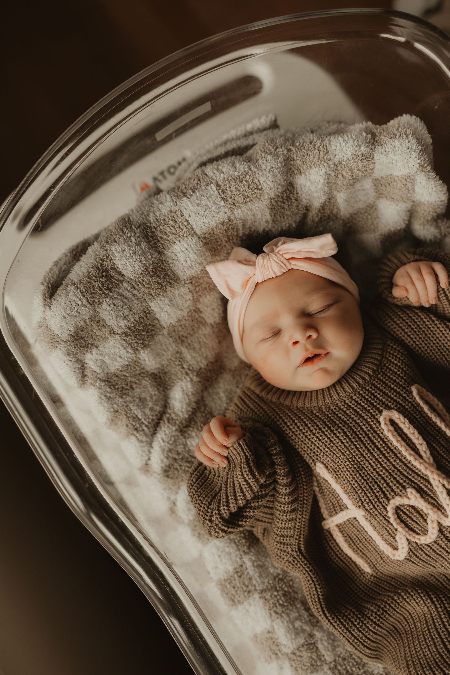 Newborn baby girl sleeping peacefully in a bassinet, wearing a knit outfit and pink bow headband on a soft blanket.