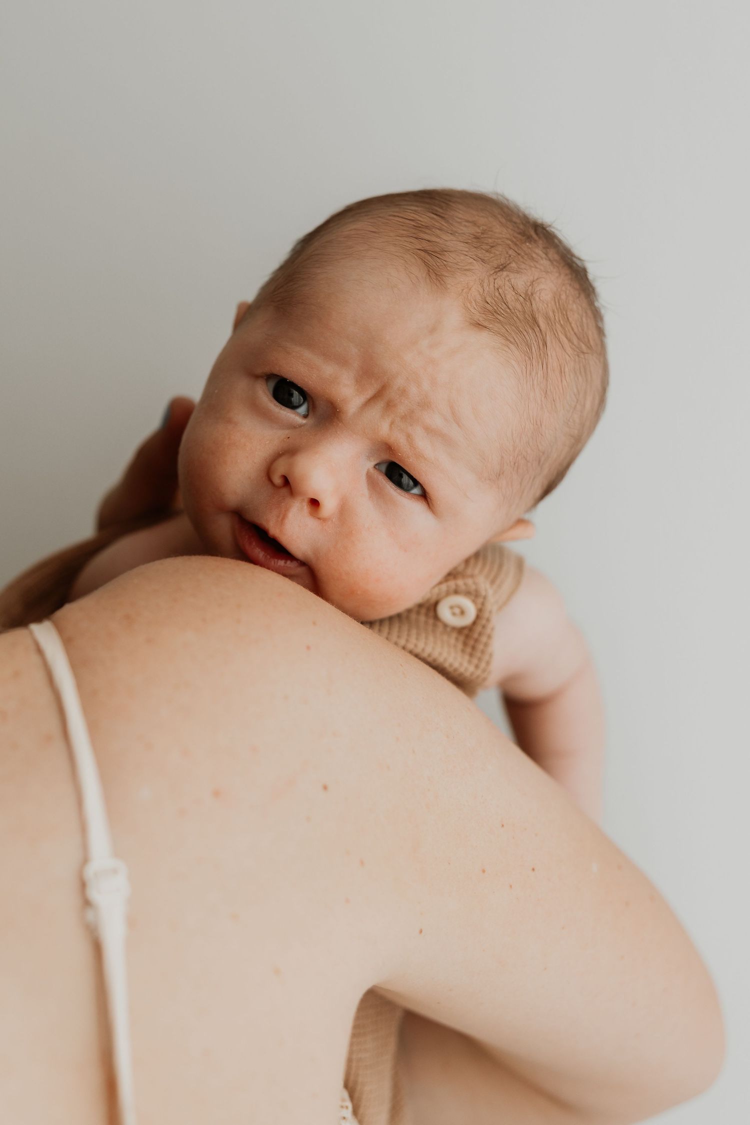 Newborn baby resting on mother's shoulder, looking up with wide eyes, wearing a neutral onesie against a light background.