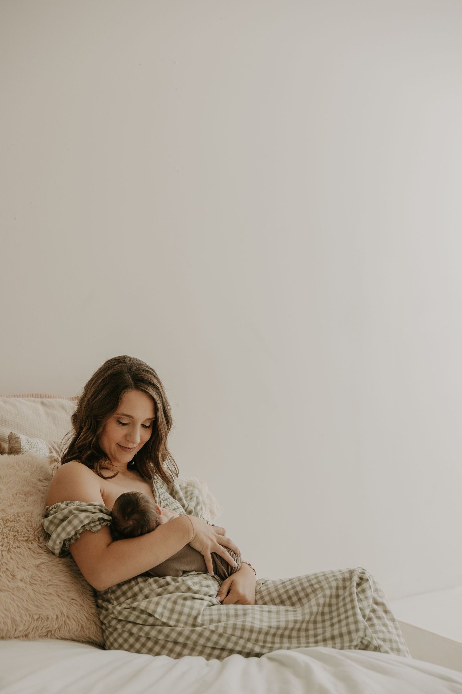 A mother tenderly breastfeeds her newborn while sitting on a bed in soft, neutral tones.