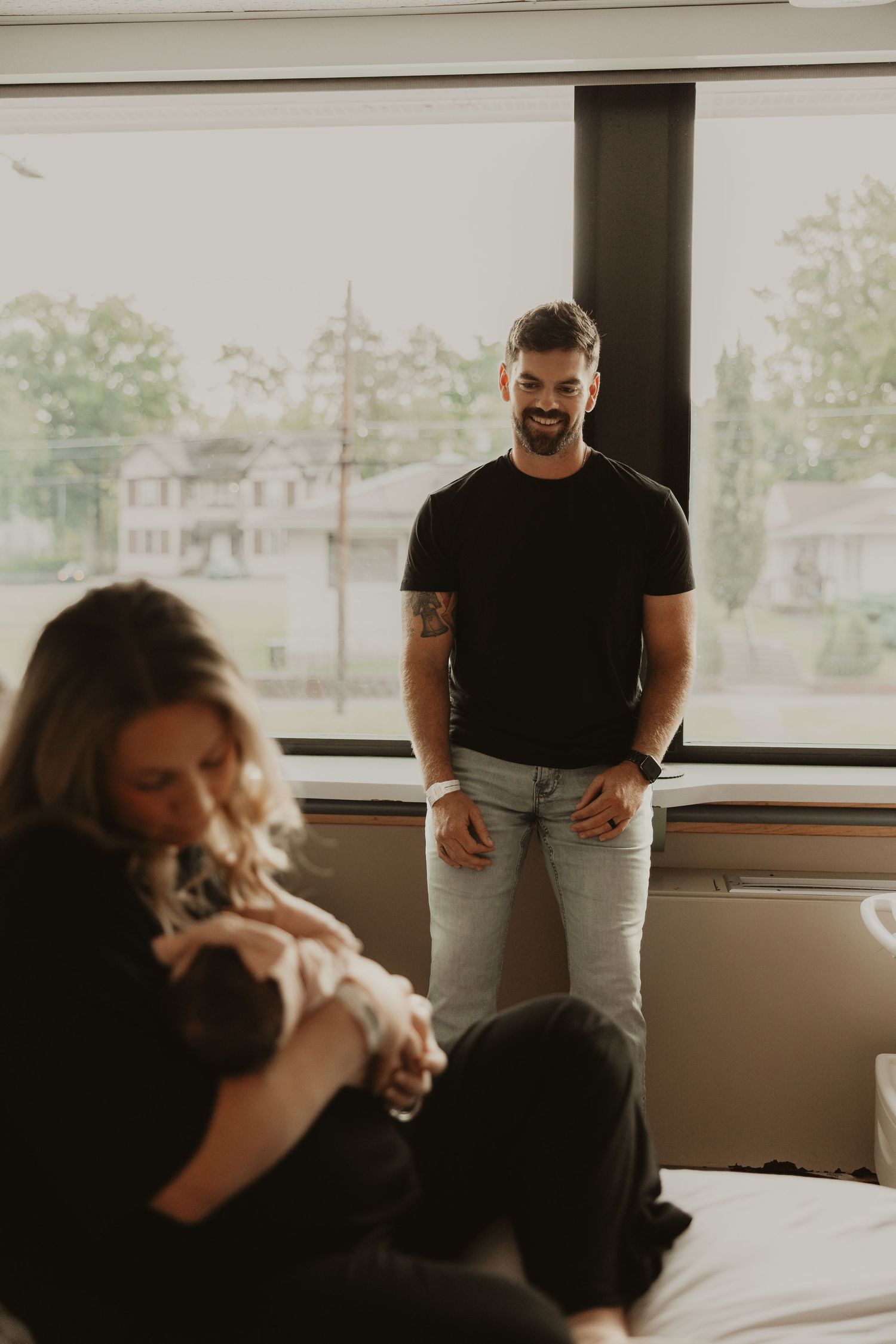 A smiling man in a black t-shirt watches lovingly as a woman holds a newborn baby in a bright hospital room.