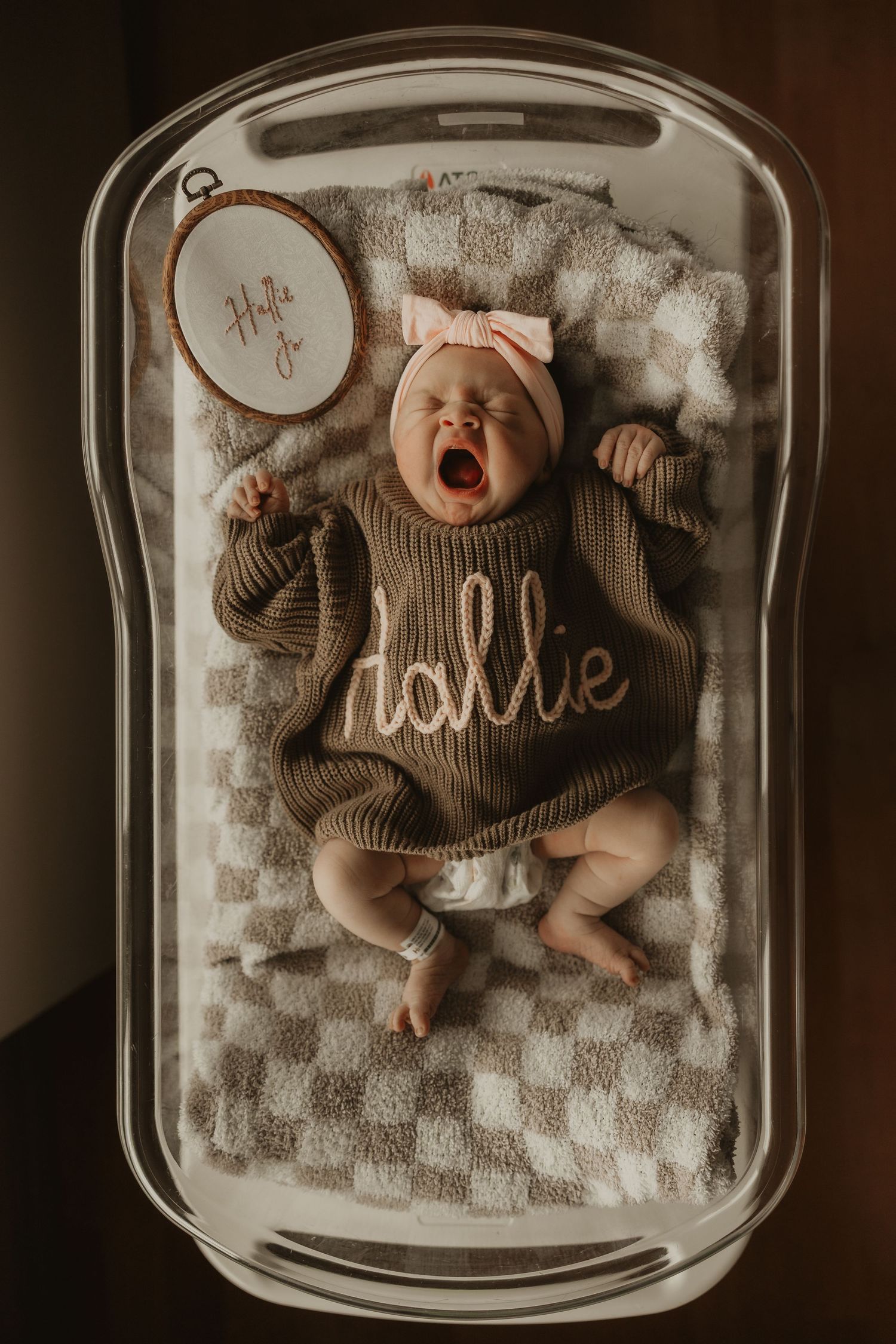 Newborn baby named Dahlie yawning in bassinet, wearing brown knit romper with name embroidered, pink bow headband.