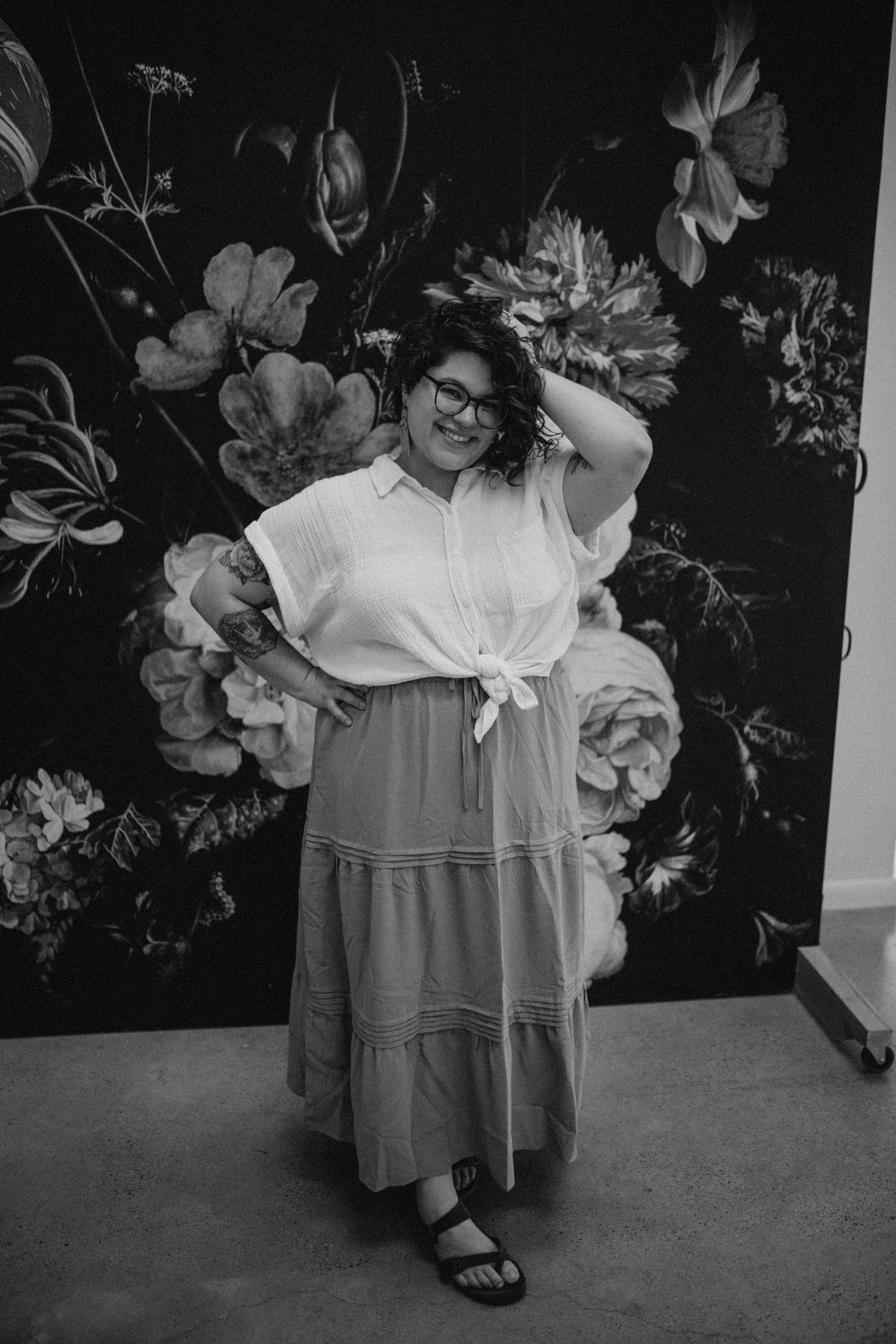 Person in glasses and dress poses against floral wallpaper in black and white.