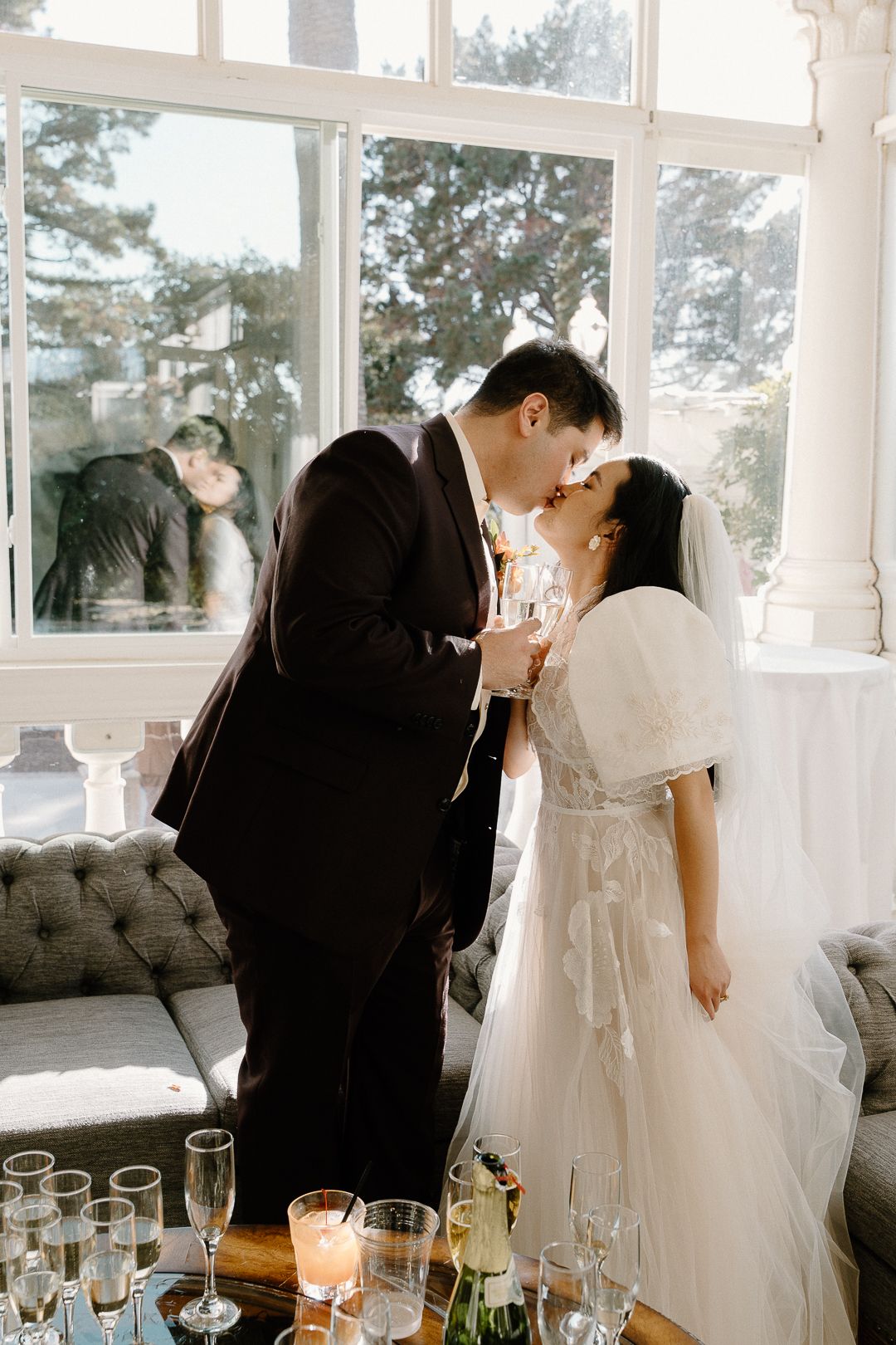 Romantic wedding couple shares intimate moments in a sunlit room with champagne glasses on a nearby table.