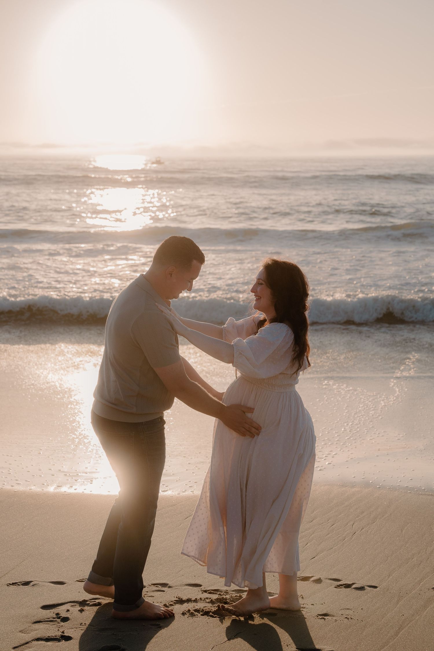 A couple dancing on beach at sunset celebrating the woman's pregnant belly as waves crash behind them at La Jolla beach in San Diego, CA.