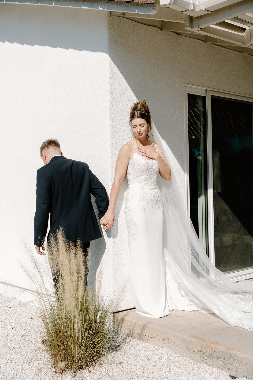 A bride in a white wedding gown and groom in black suit walk together along a white building exterior with desert grass.