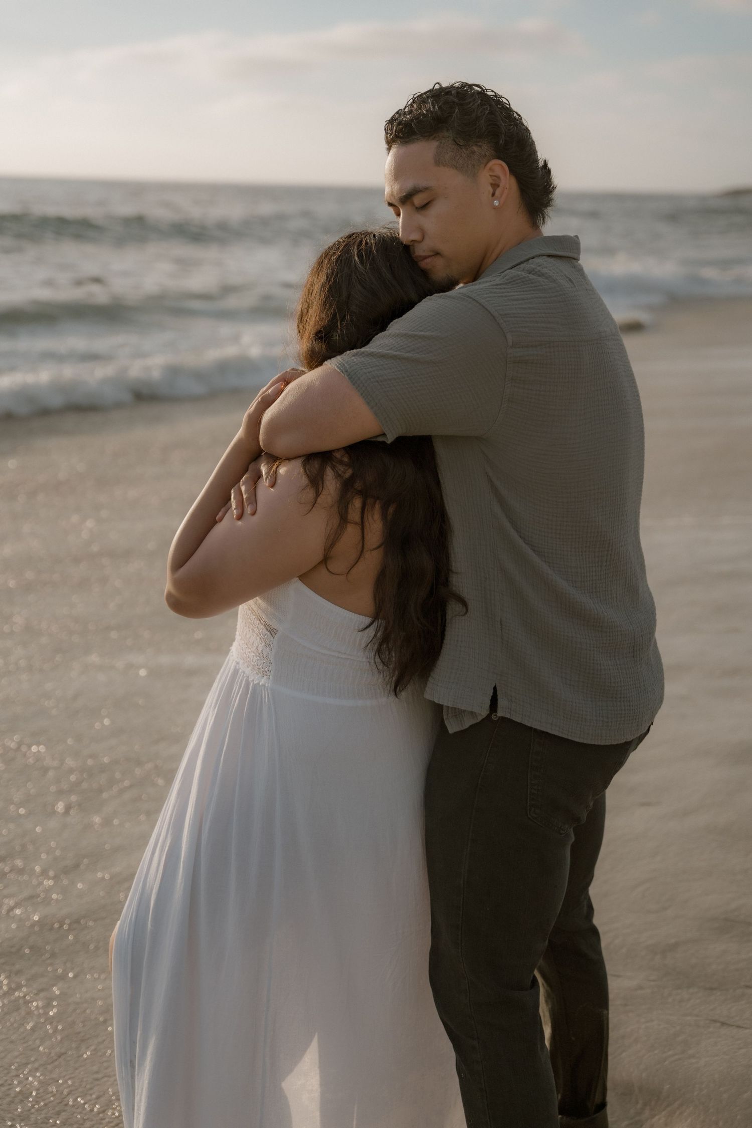 A romantic couple embraces on a beach at sunset, wearing white dress and gray shirt in La Jolla in San Diego, CA.