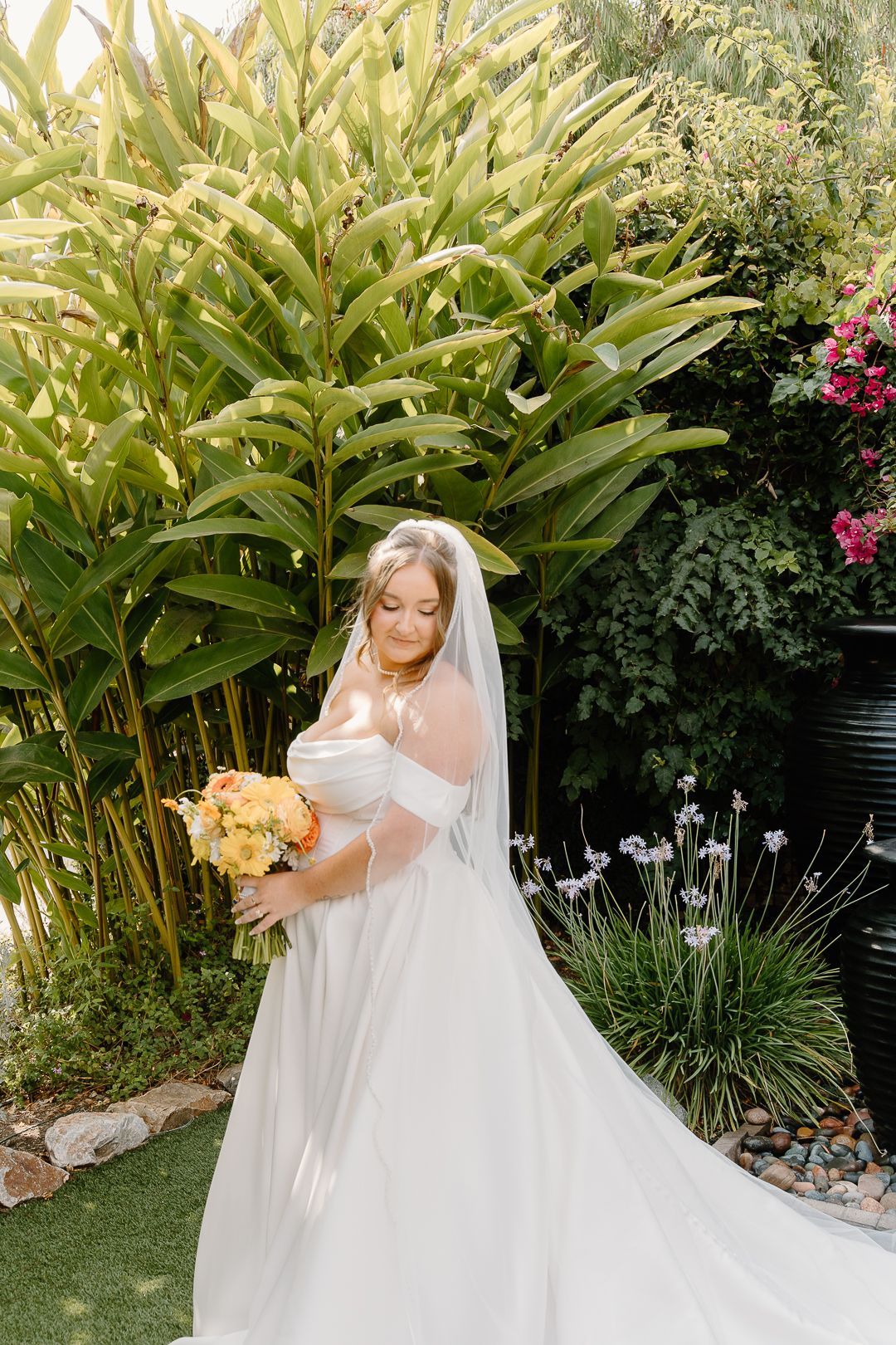 Bride in flowing white wedding dress poses gracefully in a garden setting with tall tropical plants and pink flowers.